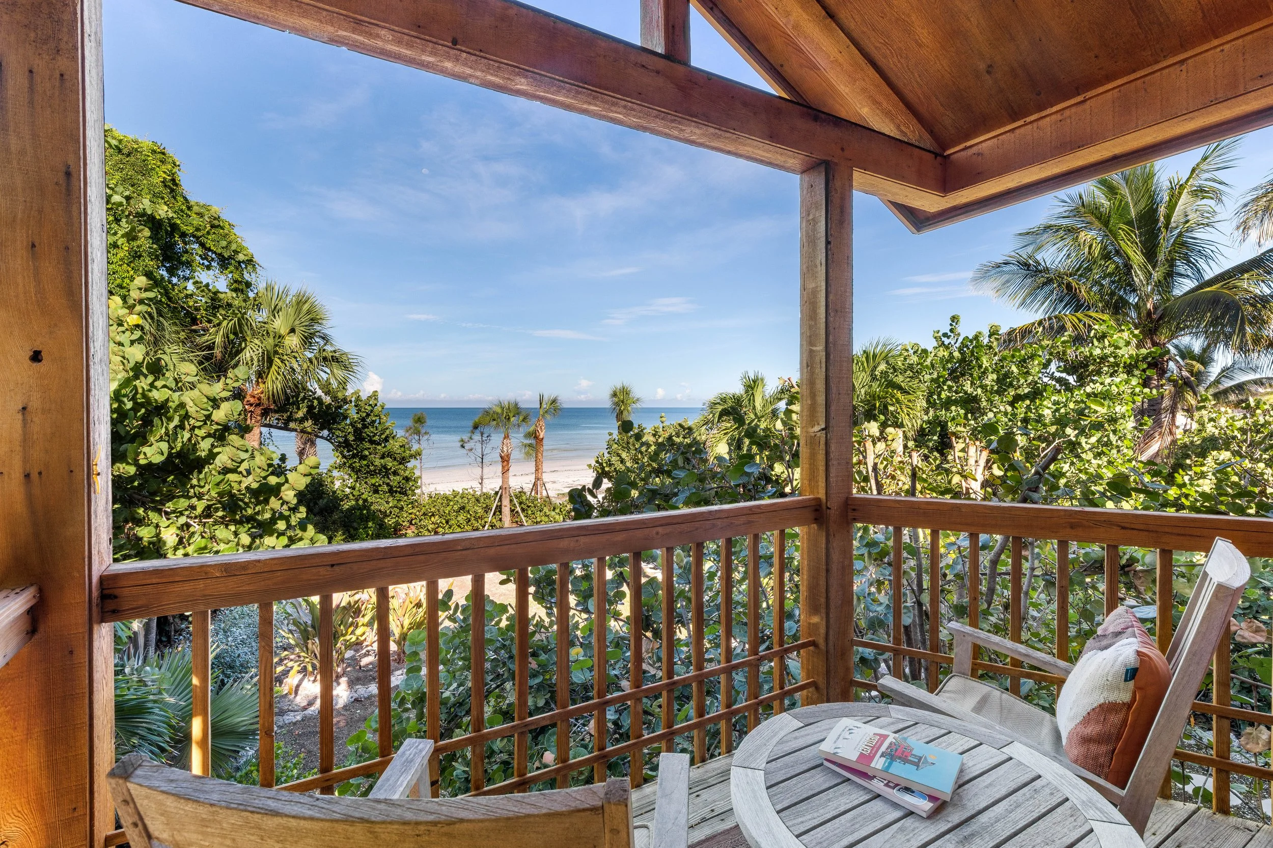 View from a wooden balcony overlooking a tropical beach with palm trees, sandy shore, and blue ocean under a partly cloudy sky.