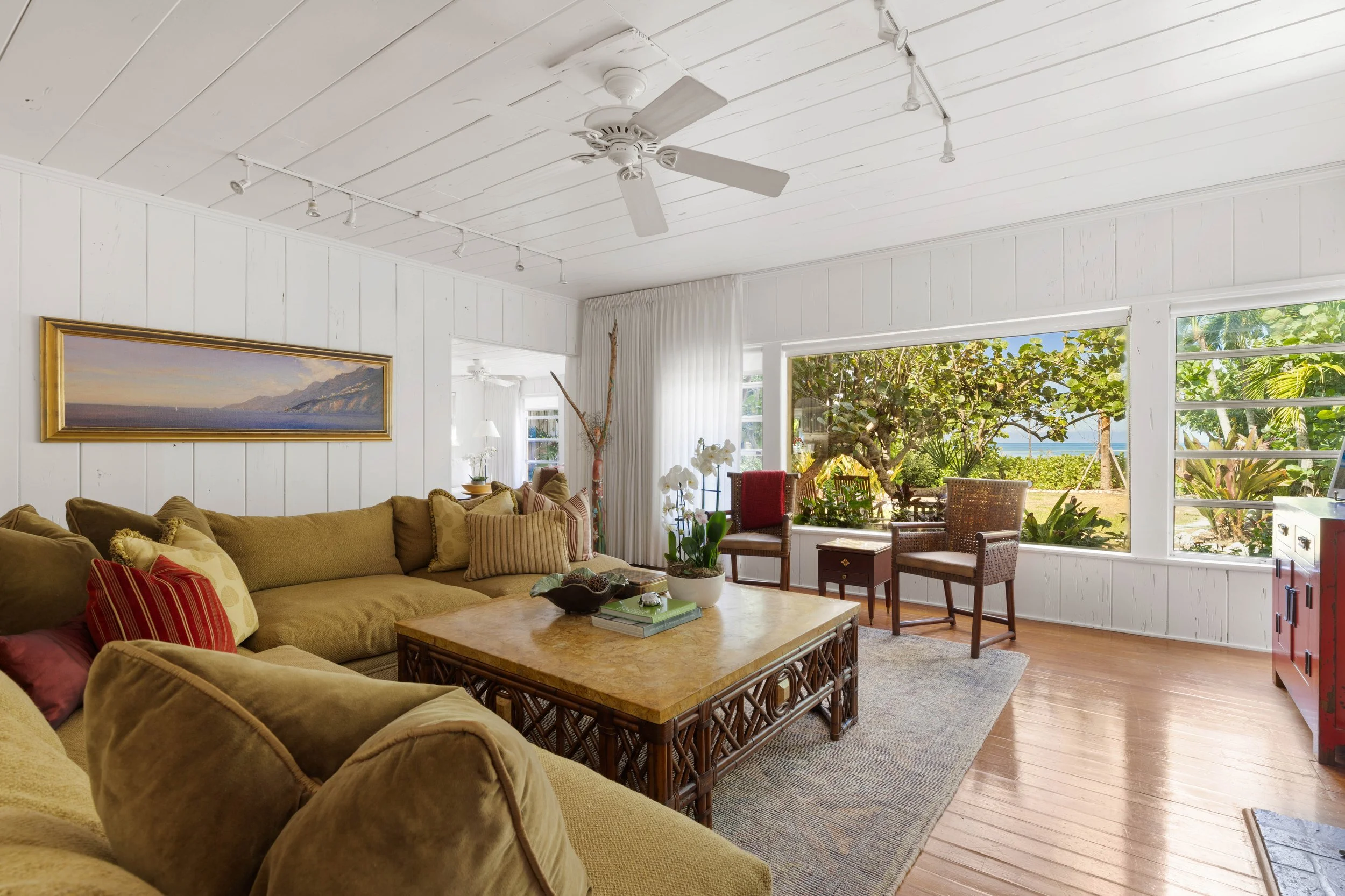 A bright, airy living room with white wooden walls and ceiling, featuring a beige sofa with red and purple pillows, a marble-top coffee table, and large windows showing a lush outdoor garden with trees and ocean in the background.