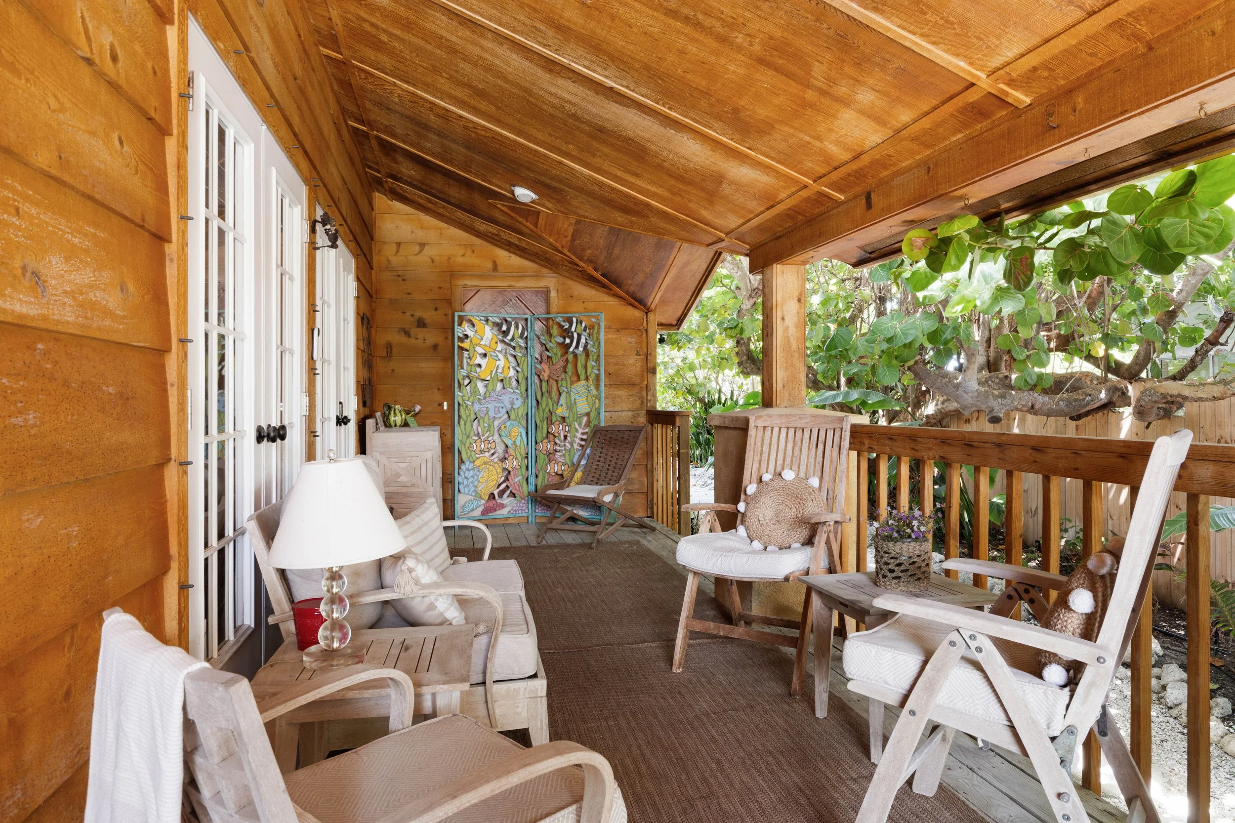 A cozy outdoor porch with wooden walls and ceiling, decorated with chairs, a table, and plants, overlooking greenery and trees.