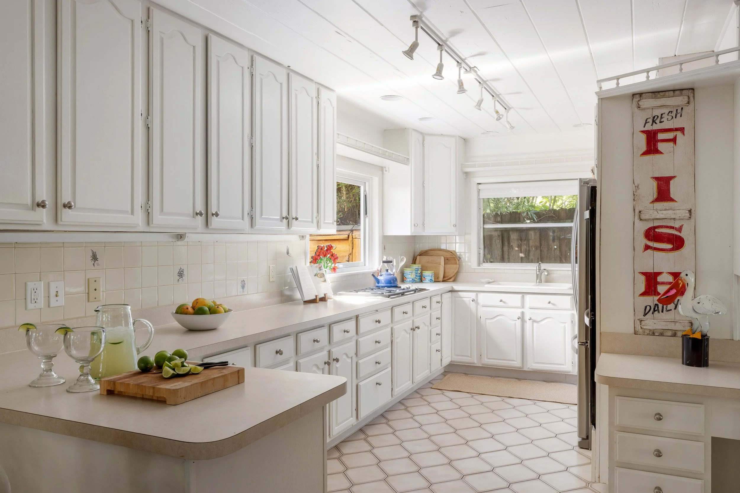 A bright, white kitchen with white cabinets and countertops. There is a window above the sink with natural light and greenery outside. The counter has a bowl of apples, a wooden cutting board, and some glasses. A vertical wooden sign with red letters reading "FRESH DAILY" is on the right side of the image.