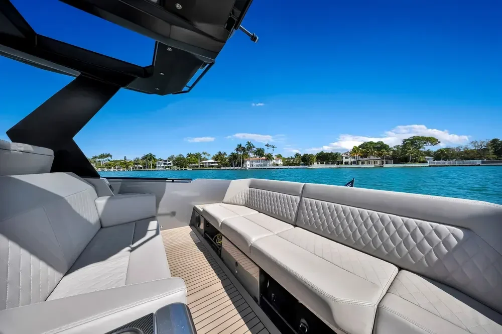 View of a yacht's deck with white cushioned seating area overlooking blue water and residential houses with palm trees on the shoreline under a clear blue sky.