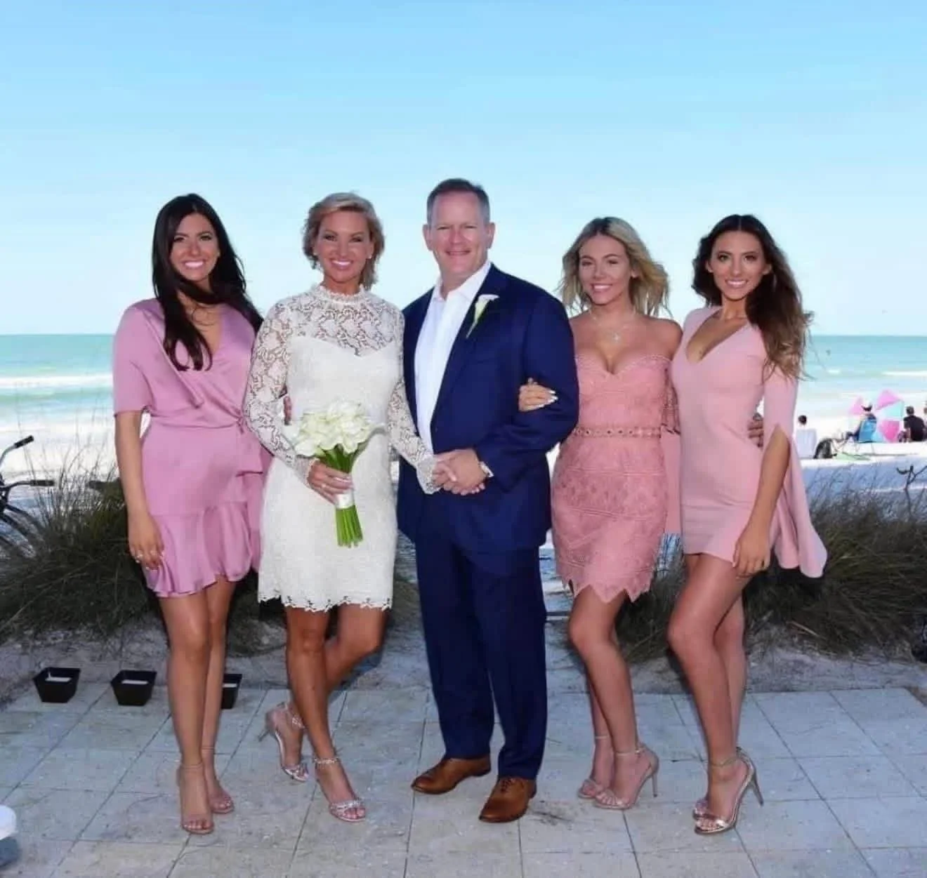 Group of five people, four women and one man, standing on a beach with ocean and sky in the background, dressed in formal attire for a wedding celebration.