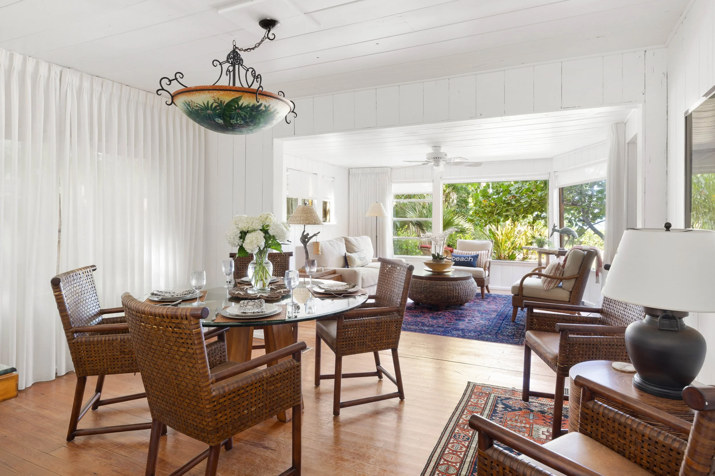 A bright, airy living and dining room with white walls, large windows showing greenery outside, wicker and upholstered furniture, a round glass dining table with chairs, and a floral centerpiece.