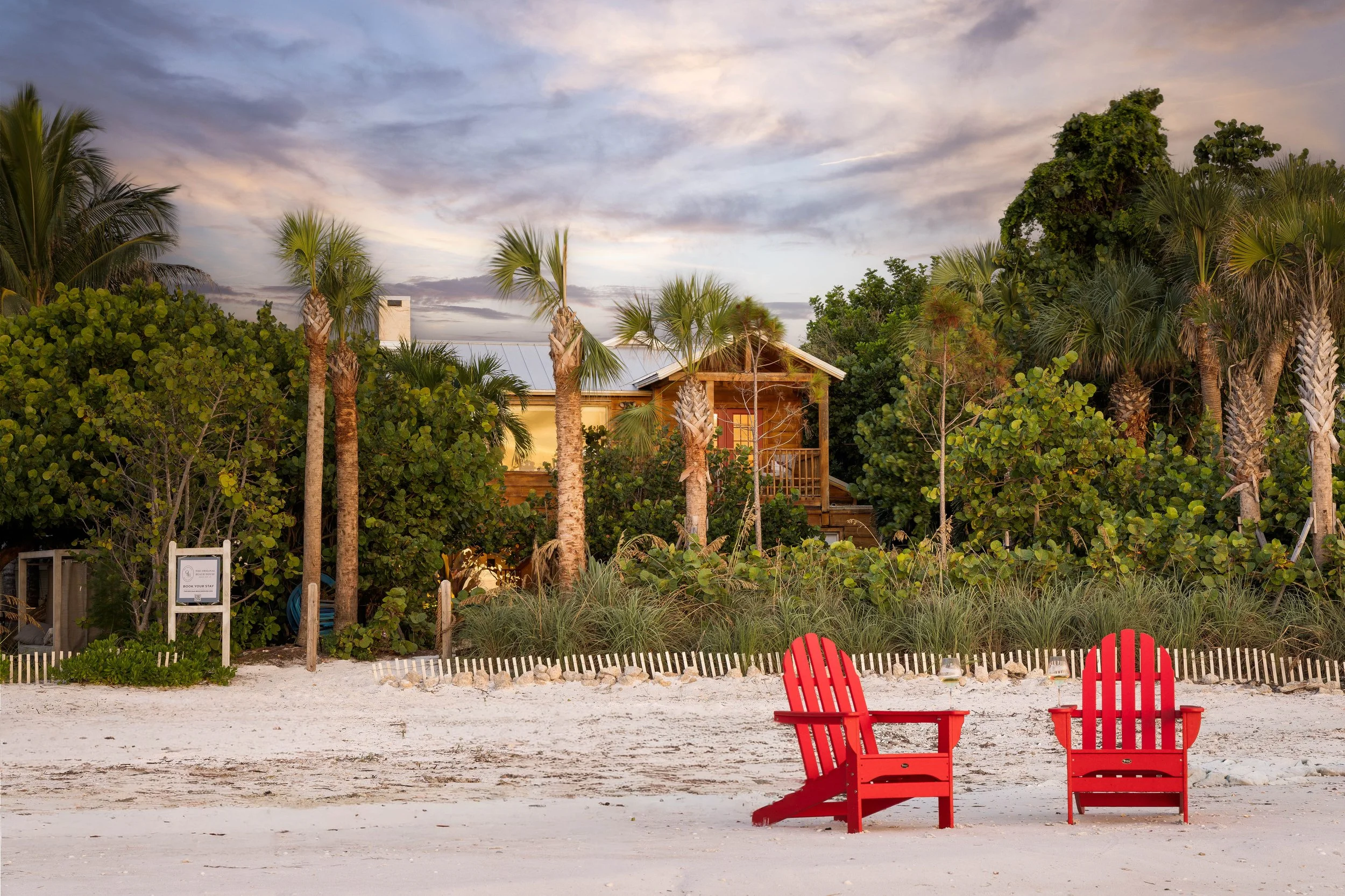 Two red Adirondack chairs on a sandy beach with a backdrop of lush greenery, trees, and a house, under a partly cloudy sky during sunset. Original Beach House, Siesta Key, Florida