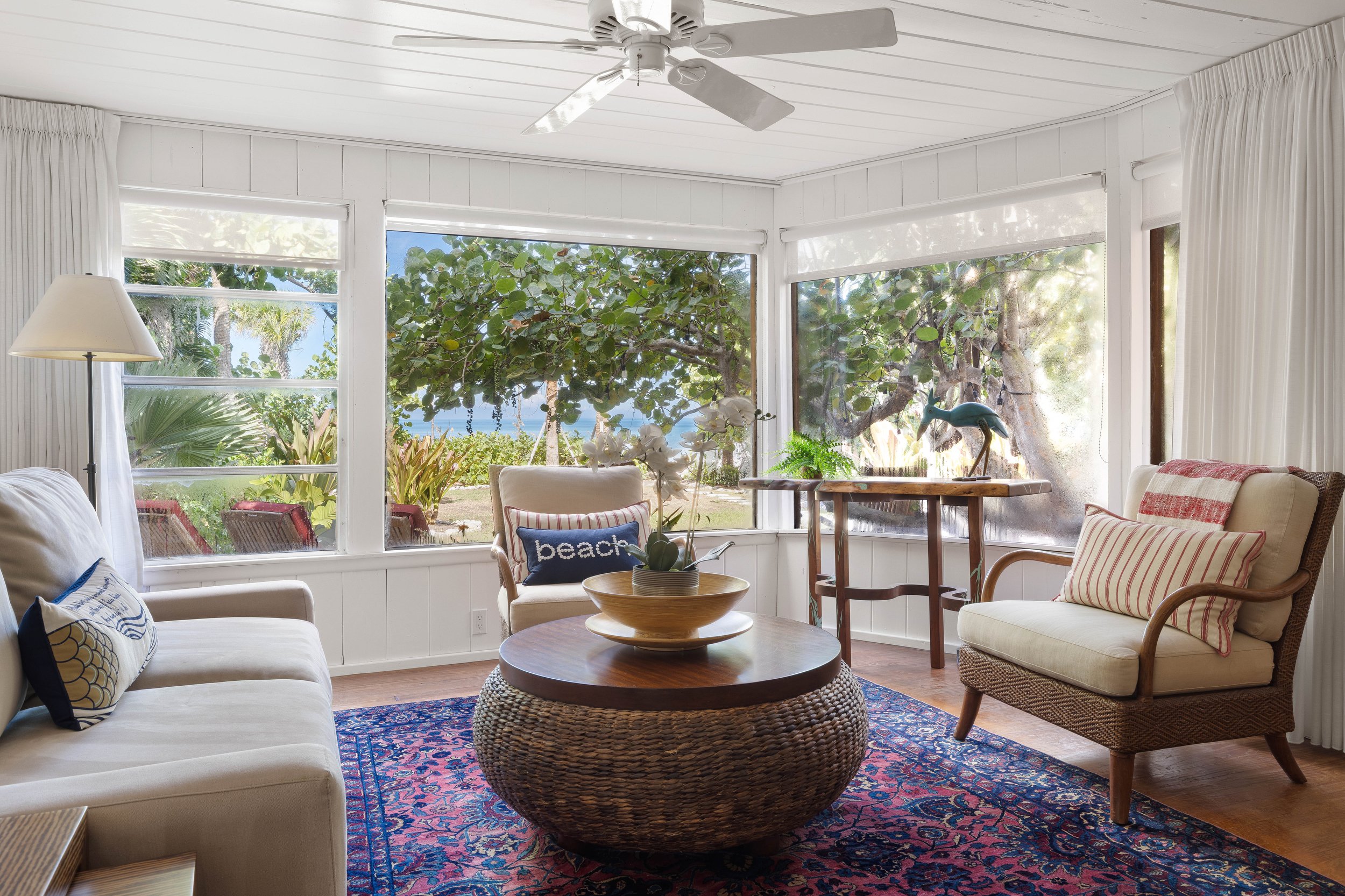 A cozy living room with a large window showing outdoor greenery and trees. The room contains a white sofa with pillows, a wicker coffee table, a wooden side table with a decorative dolphin sculpture, an armchair with a 'beach' pillow, a floor lamp, a ceiling fan, and a colorful patterned rug.