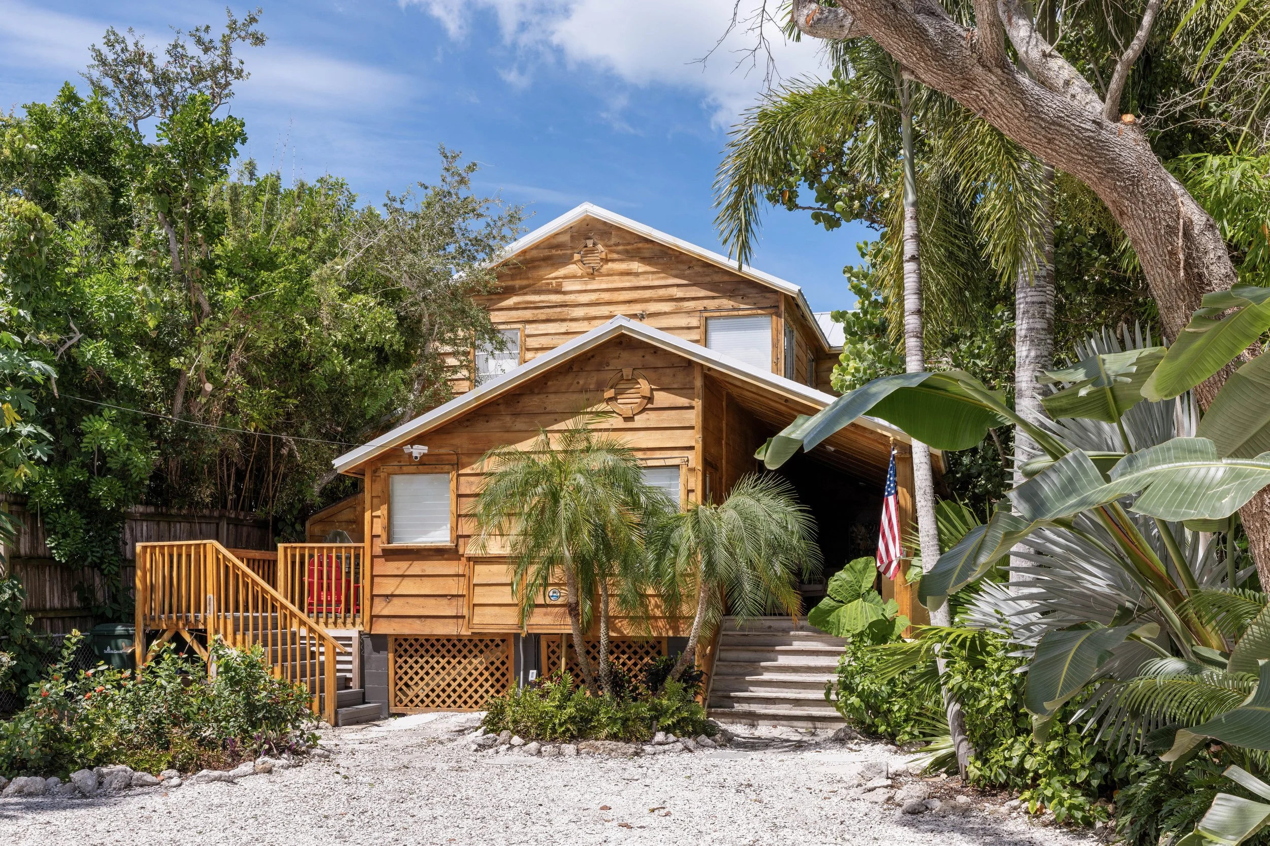 A two-story wooden house with a small porch, surrounded by lush green tropical trees and plants, under a partly cloudy sky. Original Beach House, Siesta Key, Florida