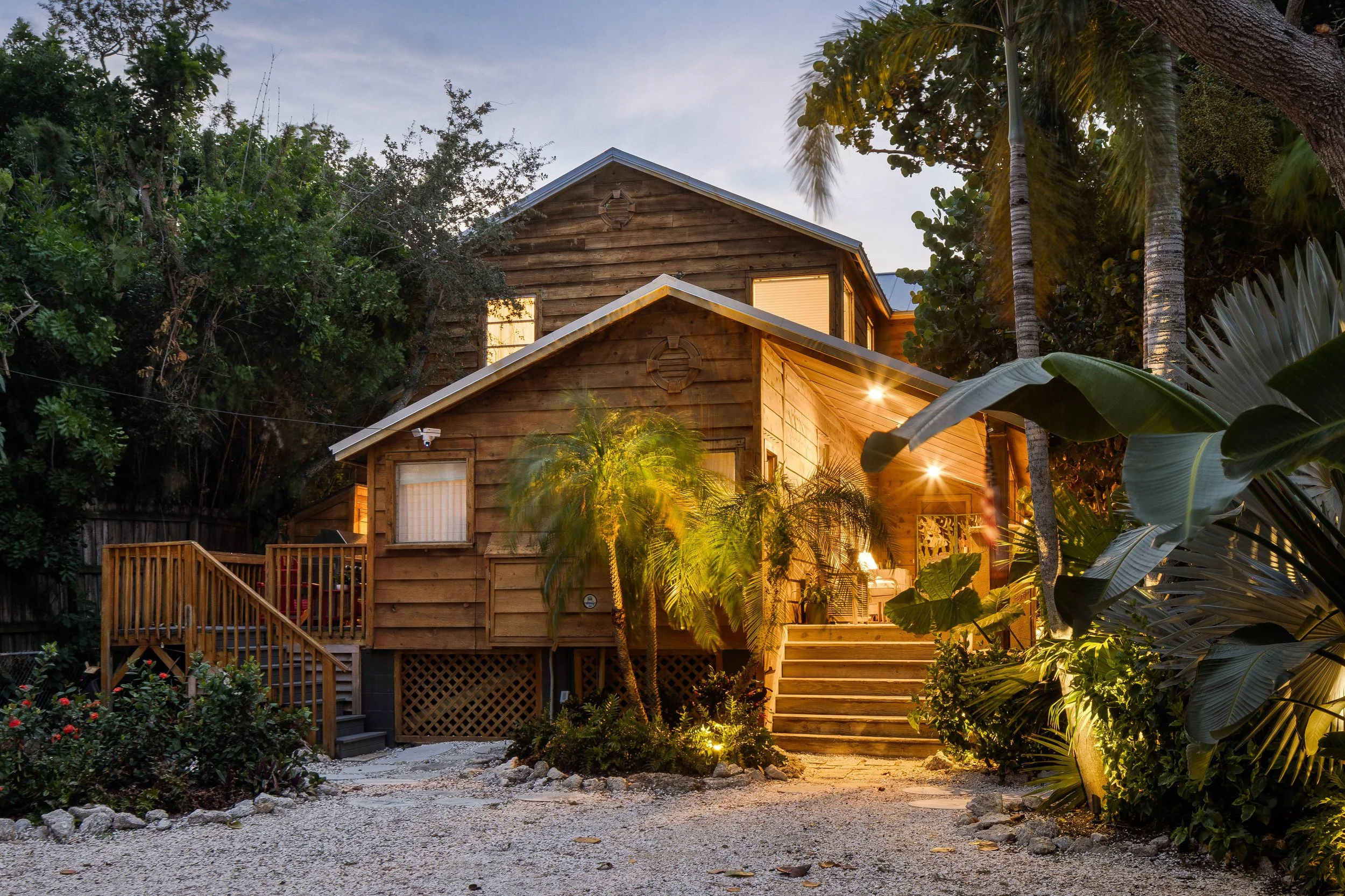 Wooden house in a lush garden with trees and plants, illuminated by outdoor lighting at dusk or early evening.