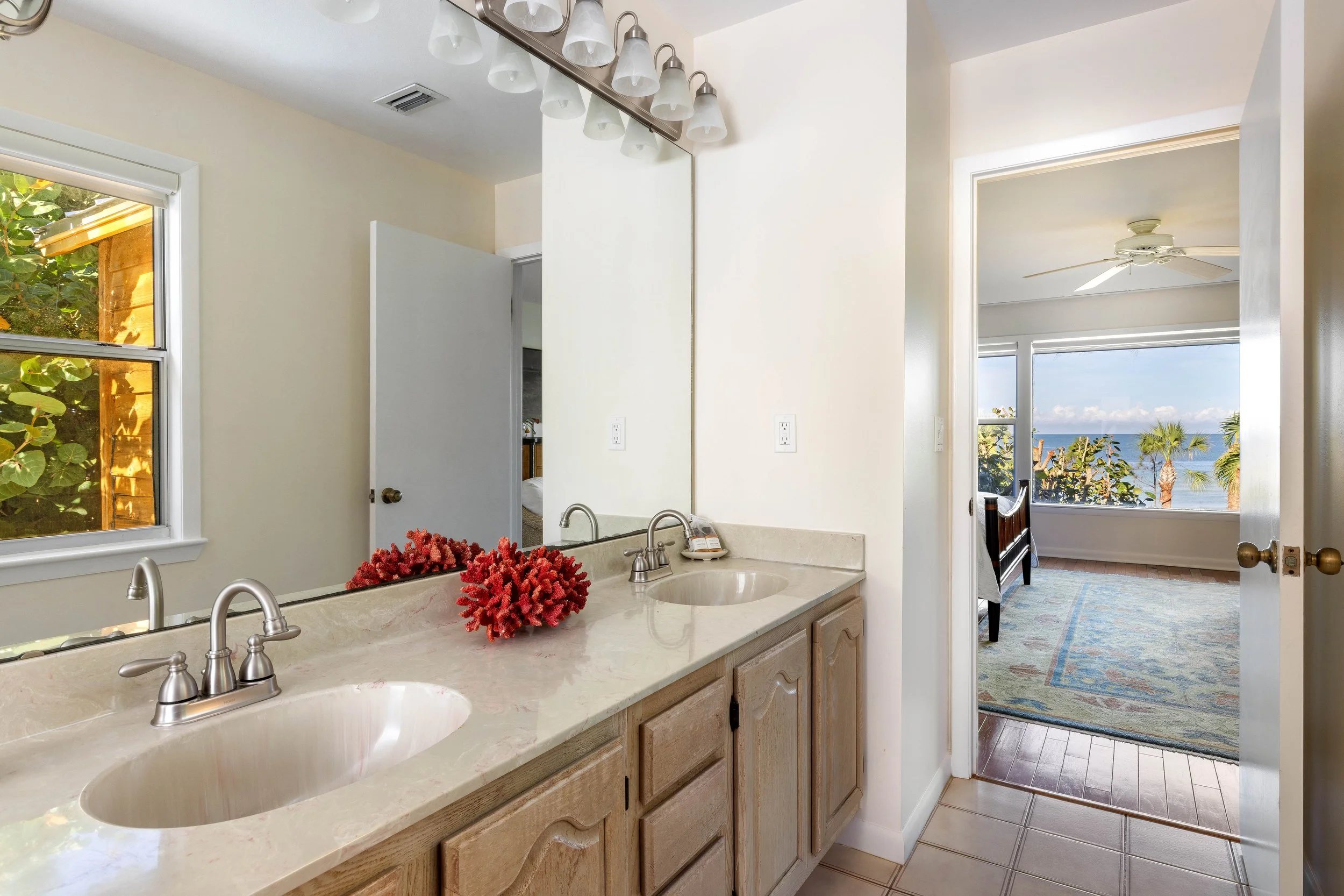 A bathroom with a double sink vanity, a mirror, and a window revealing a view of the beach and palm trees. There is a ceiling fan in the adjoining room.
