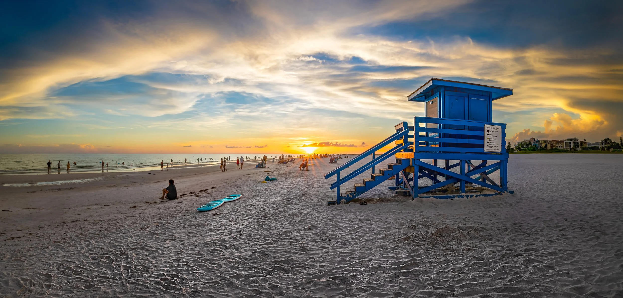 Sunset over a beach with people walking and sitting on the sand, a blue lifeguard tower, and a paddleboard on the ground.