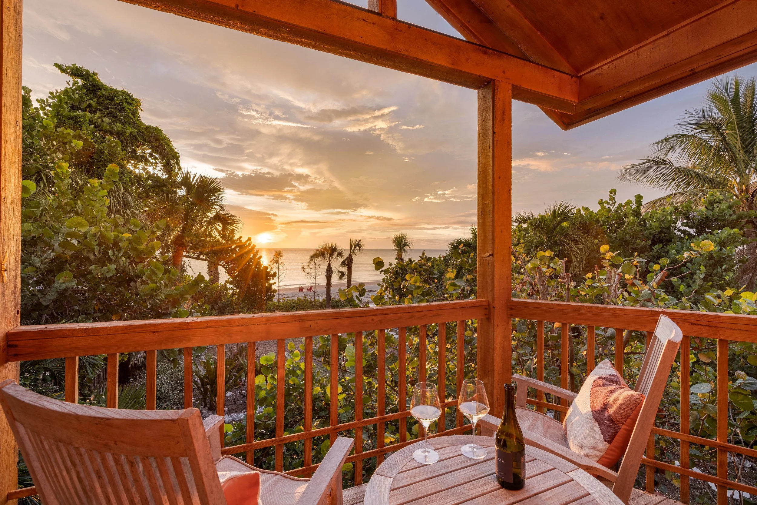 A wooden balcony overlooking a beach with palm trees during sunset, with glasses of wine and a bottle on a table. Original Beach House, Siesta Key, Florida