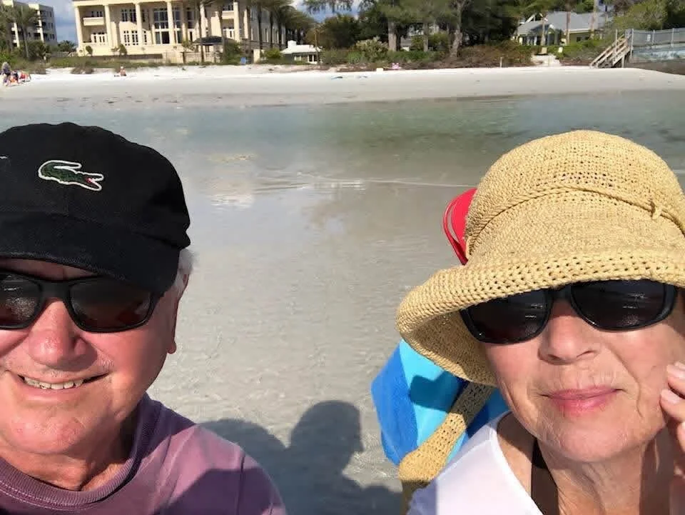 A smiling older man and woman wearing sunglasses on a beach with white sand, in front of houses and trees, with clear water and blue sky in the background.