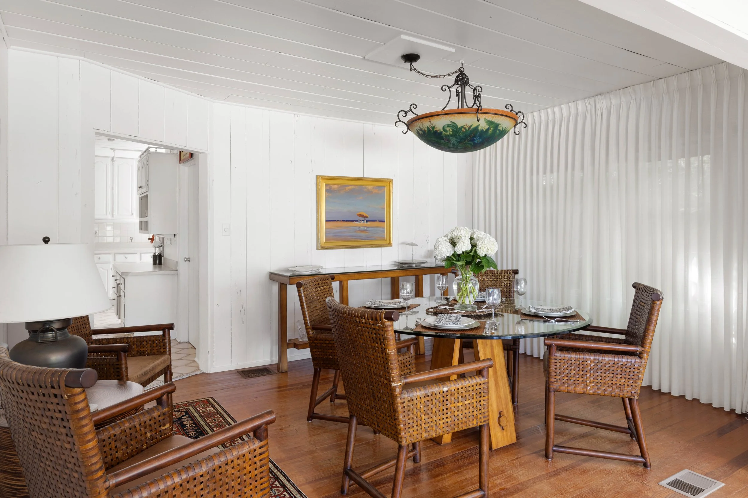 A dining room with a round glass table set for six, wicker chairs, white walls, a painting of a beach scene, and a floral centerpiece, with a decorative ceiling light and a view into the kitchen.