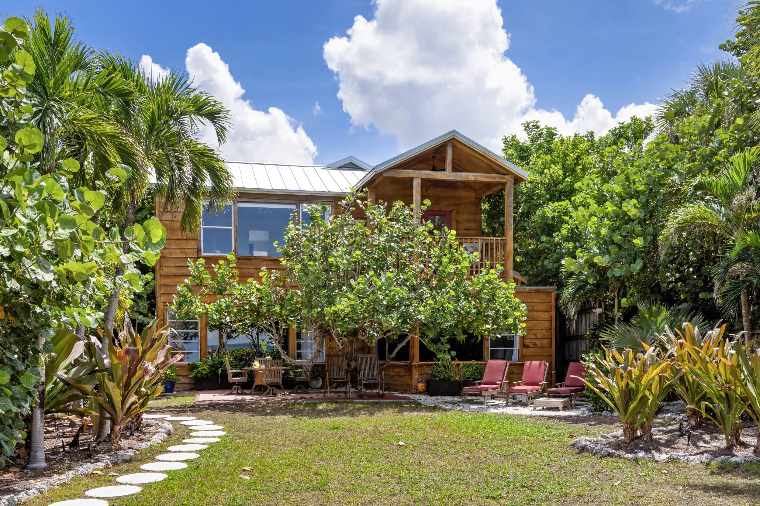 A wooden house with a small balcony, surrounded by lush green plants and trees, with patio chairs and a stone pathway in the backyard under a partly cloudy sky. Original Beach House, Siesta Key, Florida
