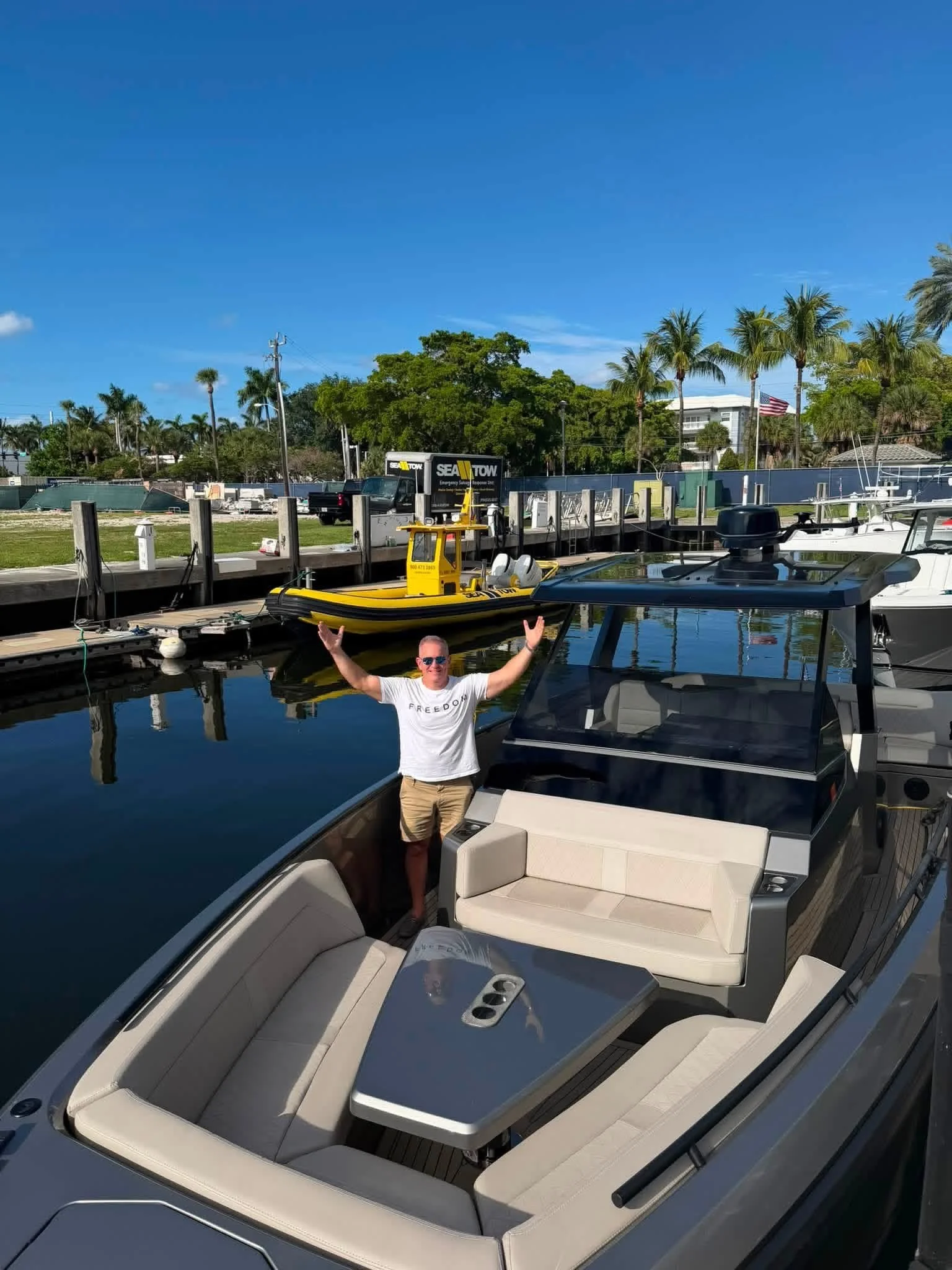 A man standing on a boat dock with arms raised, smiling, in a marina with boats, palm trees, and a clear blue sky in the background.