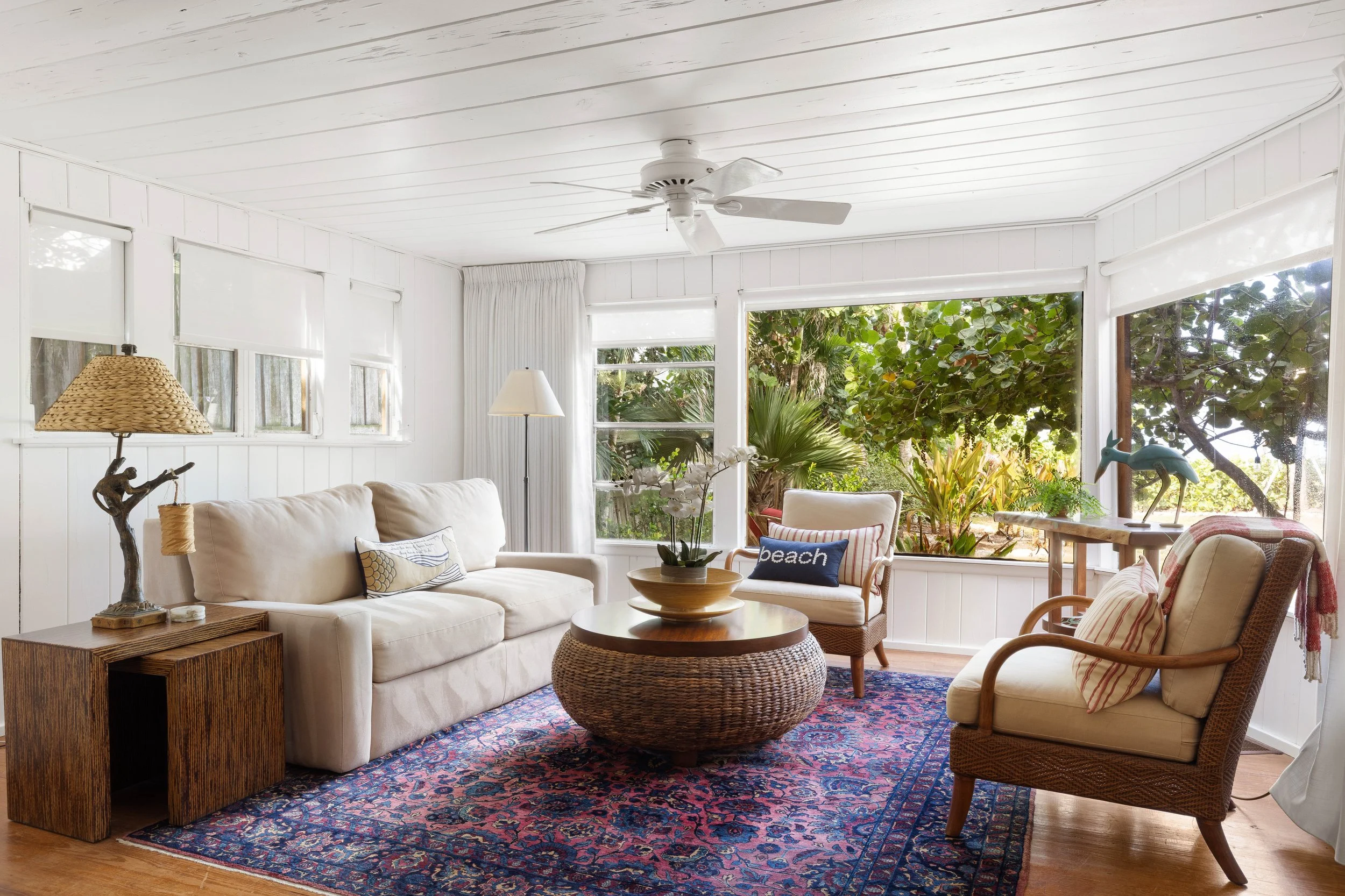 A sunlit living room with white walls and large windows showing lush greenery outside. The room features a beige sofa, wicker chairs with striped and plain cushions, a round woven coffee table, and a colorful Persian rug. Decor includes a table lamp 
