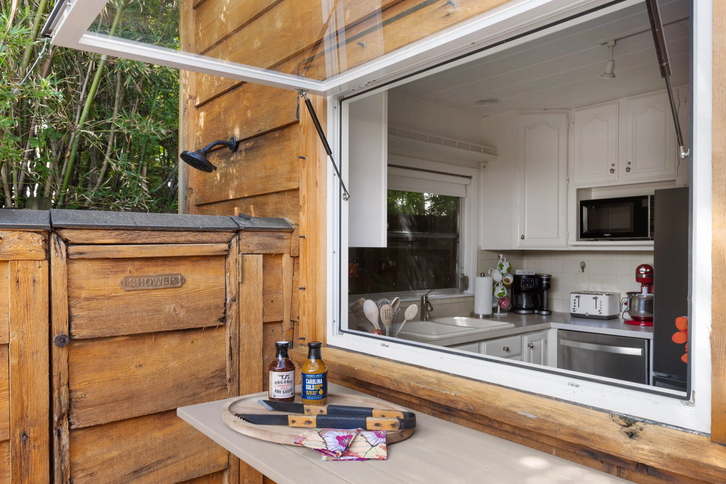 Wooden outdoor kitchen with open window showing a kitchen interior. Counter with utensils and two bottles of barbecue sauce outside. Shower labeled wooden cabinet to the left. Showerhead attached to the wood wall above the counter. Forest plants behi