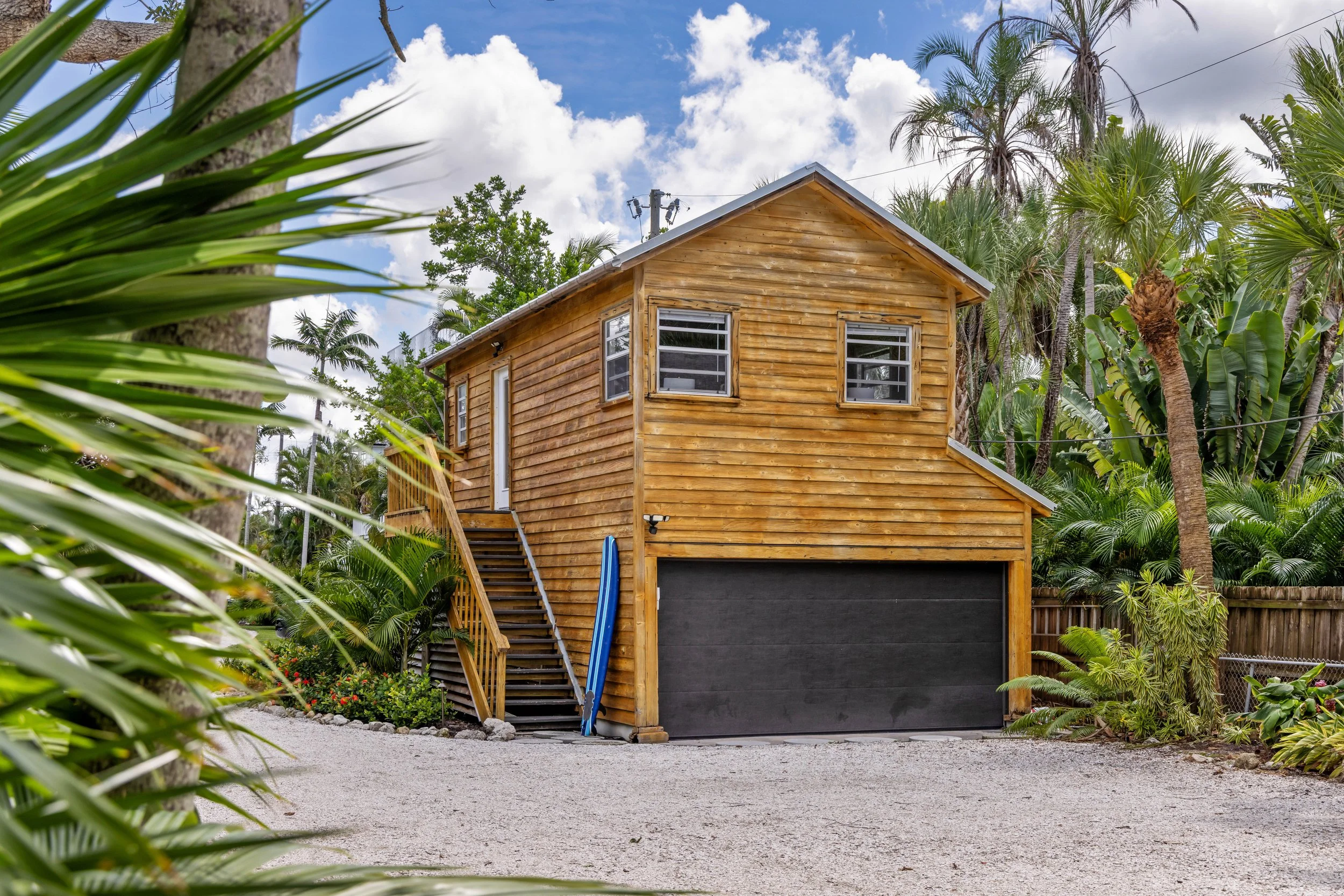 A two-story wooden house with a black garage door in a lush tropical setting, surrounded by tall palm trees and green foliage under a partly cloudy blue sky on Siesta Key.