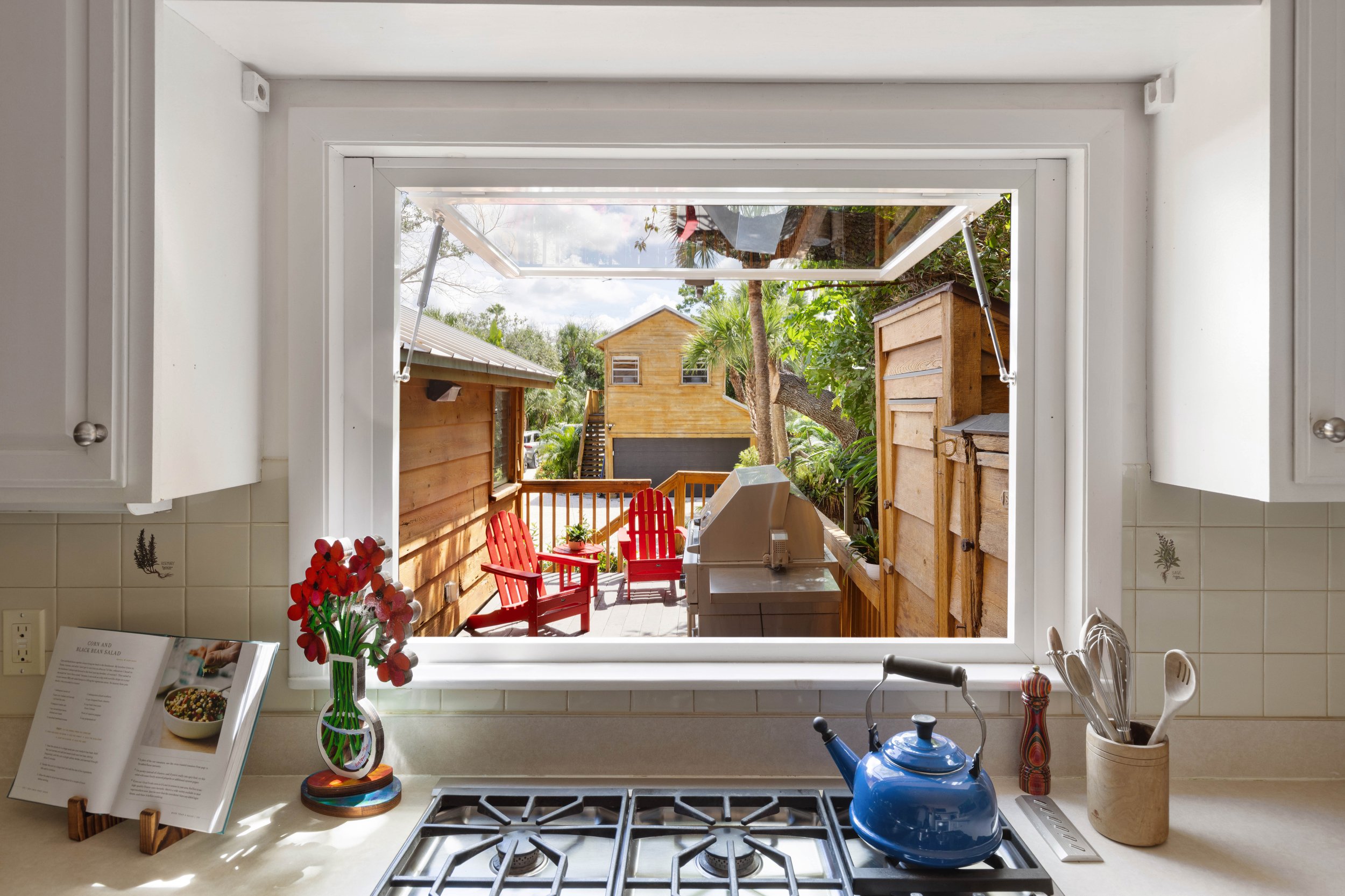 View from inside a kitchen looking out to a backyard through an open window. The countertop has a blue kettle, a vase with red flowers, a cookbook, and kitchen utensils. The backyard features a wooden deck with two red chairs, green plants, trees, and a yellow house with a black garage door.