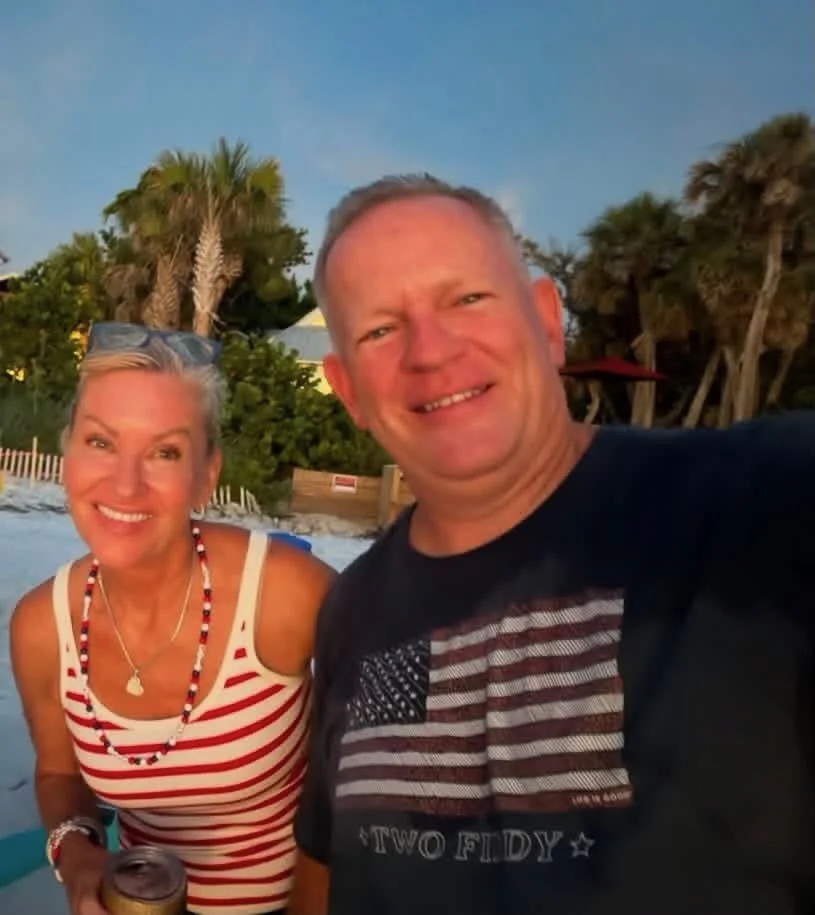 A smiling couple taking a selfie on a beach with palm trees and a clear sky in the background
