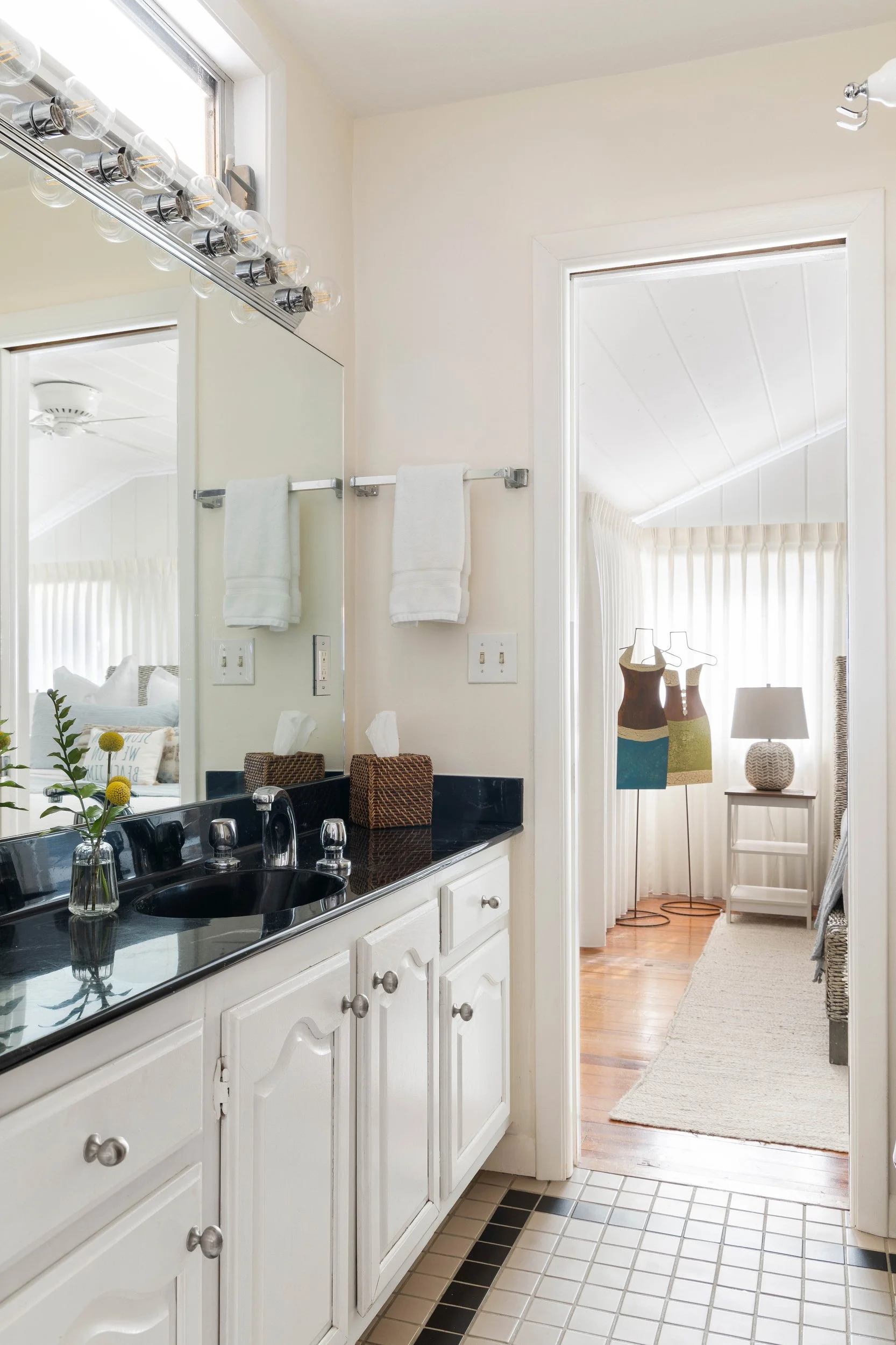 A bathroom with white cabinetry, black countertop, and a black sink. A small vase with yellow flowers is on the counter. A large mirror with exposed light bulbs hangs above the sink. Towels are hanging on a bar beside the mirror. Adjacent to the bathroom is a room with three mannequin busts, a table lamp, and a window with curtains.