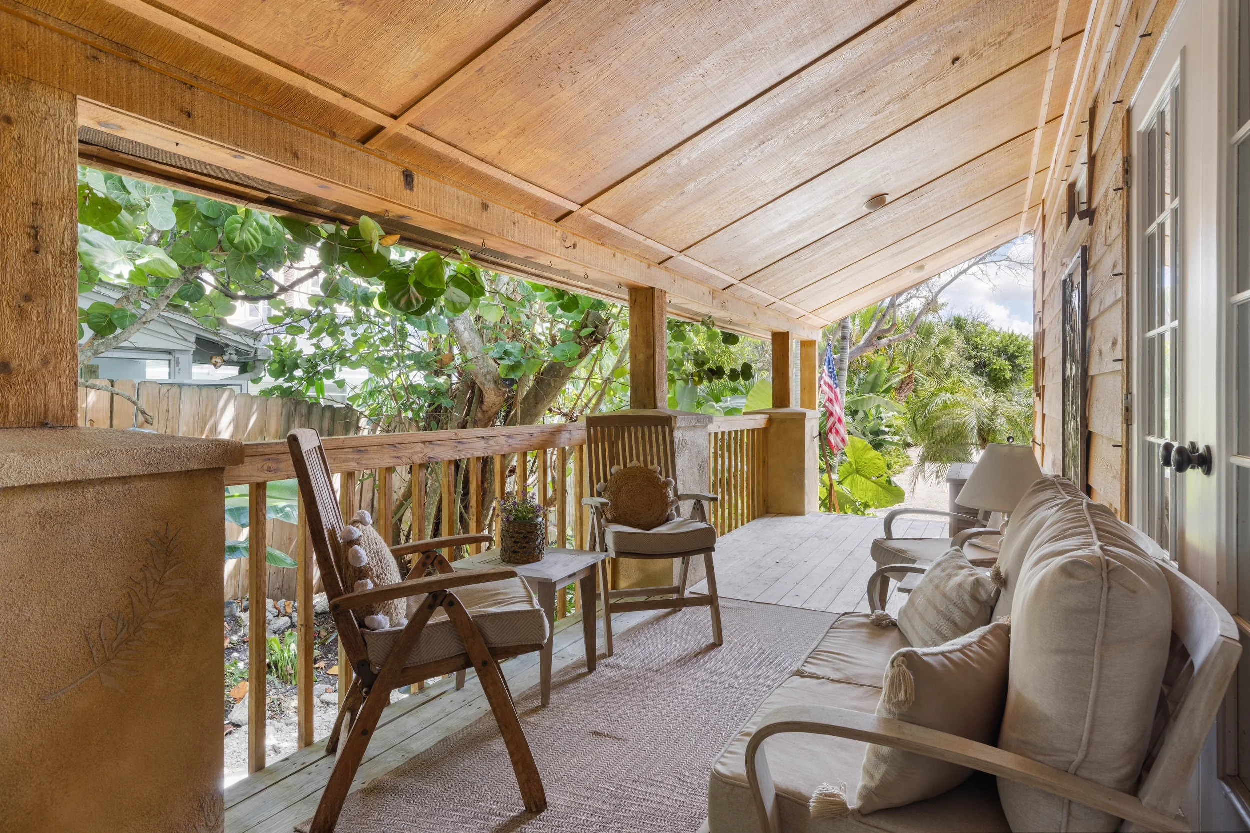 Covered outdoor porch with chairs, a sofa, potted plant, and views of trees and greenery outside.