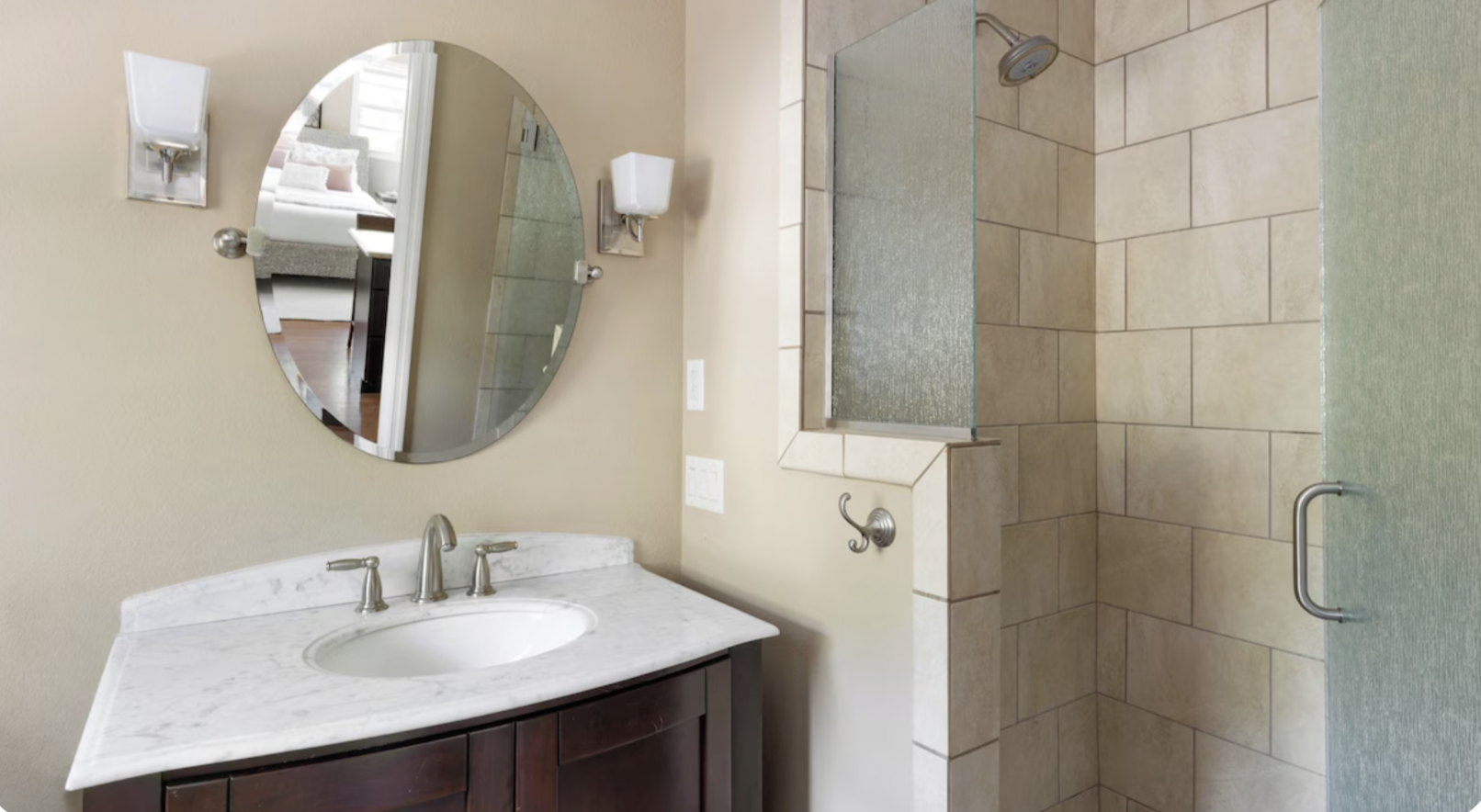 Bathroom with a white marble sink vanity, oval mirror, wall-mounted lights, and a glass-enclosed shower area with beige tiles and a handle.