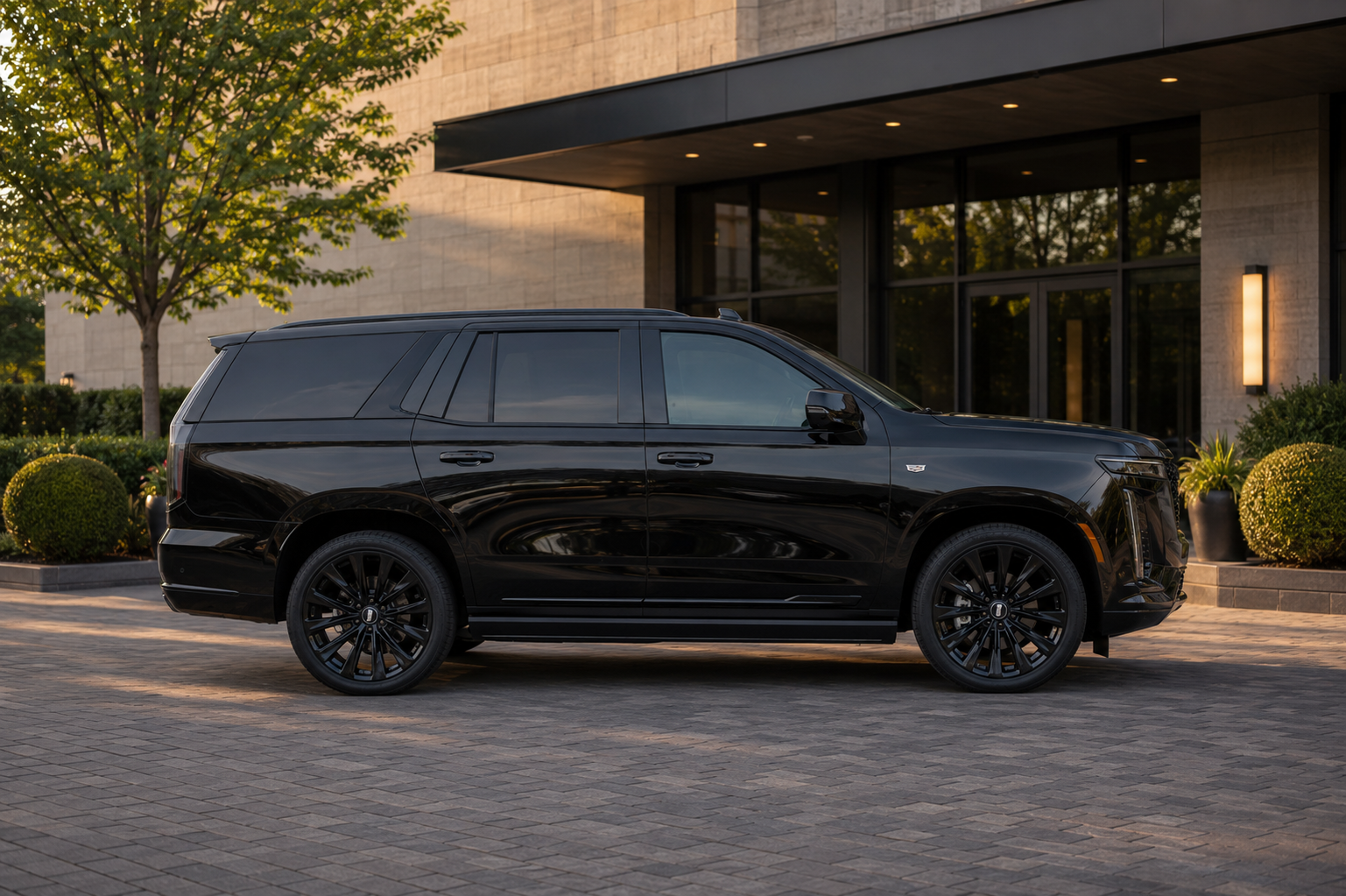 A black luxury SUV parked in front of a modern building with large glass doors, surrounded by landscaped bushes and trees during sunset.