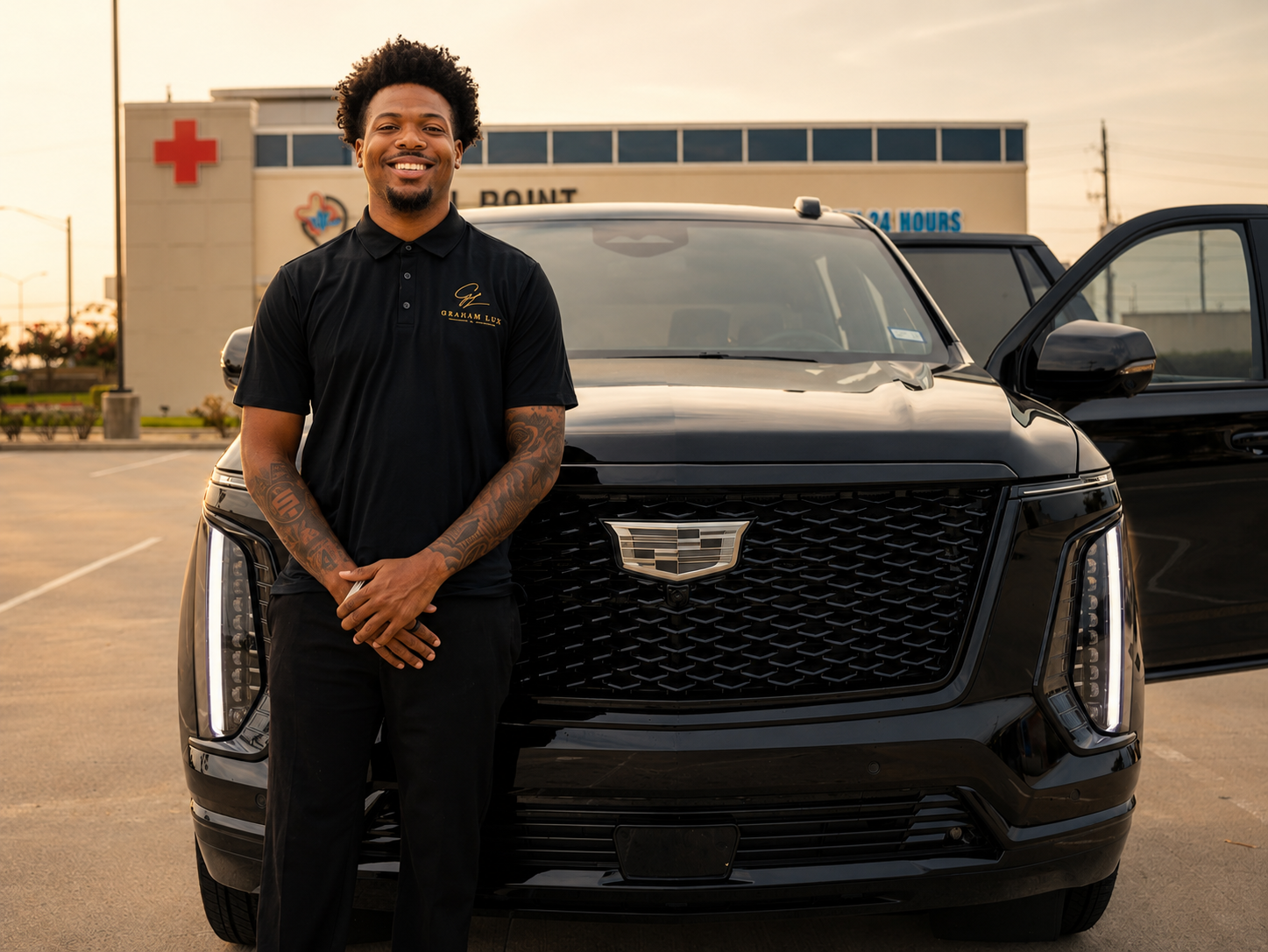 A smiling man with tattoos wearing a black polo shirt with gold embroidery, standing in front of a shiny black Cadillac SUV in a parking lot near a medical center with a red cross sign, during sunset.