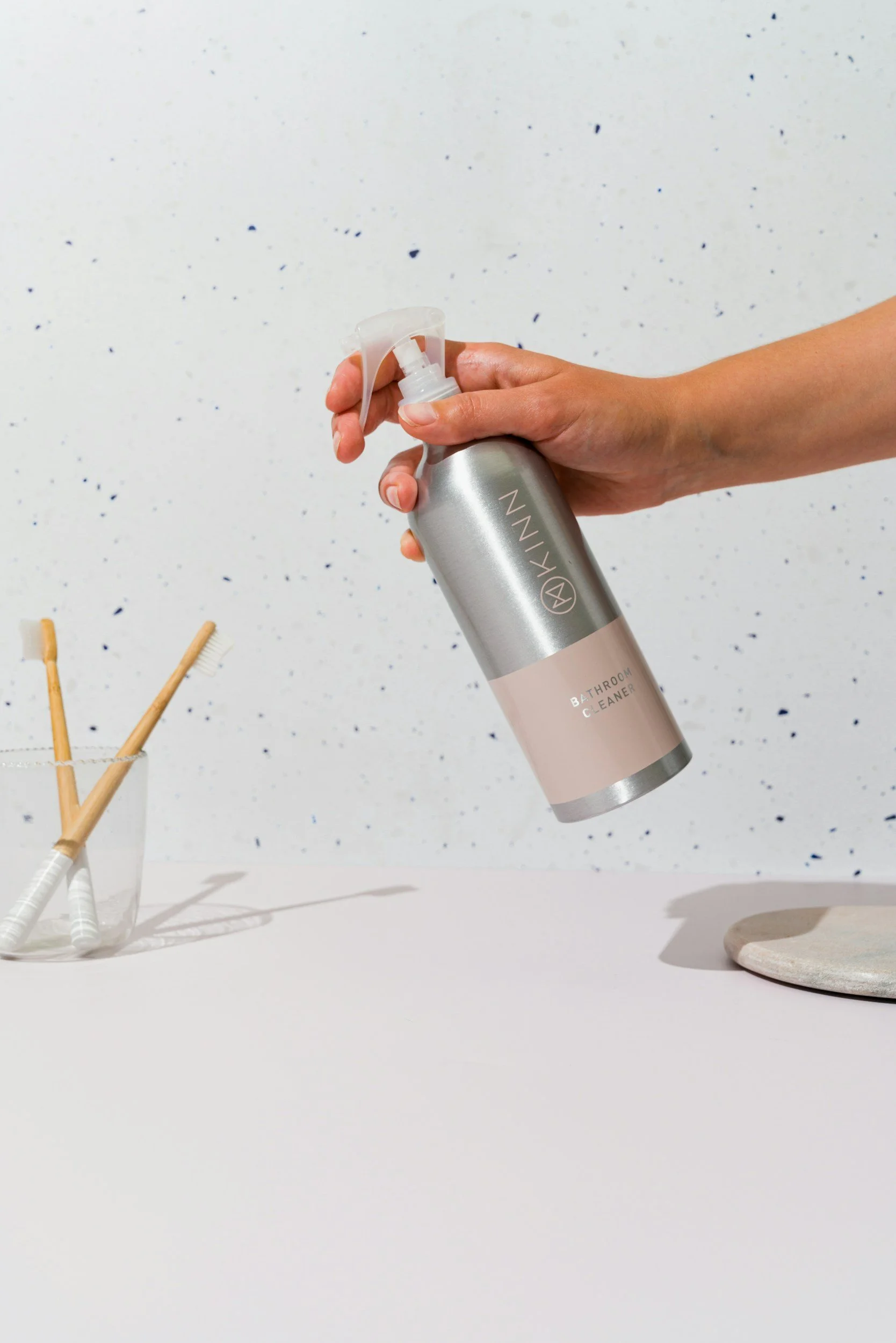 Hand holding a gray and beige spray bottle labeled "Bathroom Cleaner" over a white countertop with toothbrushes in a glass and a small round stone.