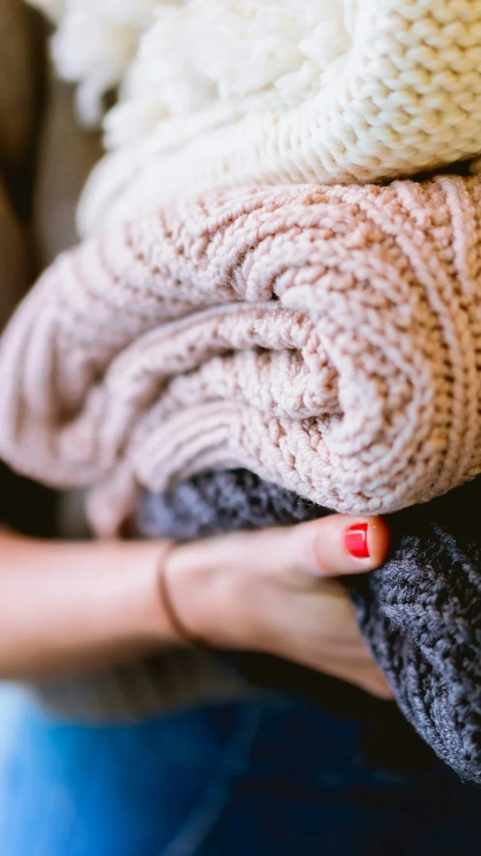 Close-up of a person holding a stack of rolled, thick knitted blankets in beige, pink, gray, and black colors.