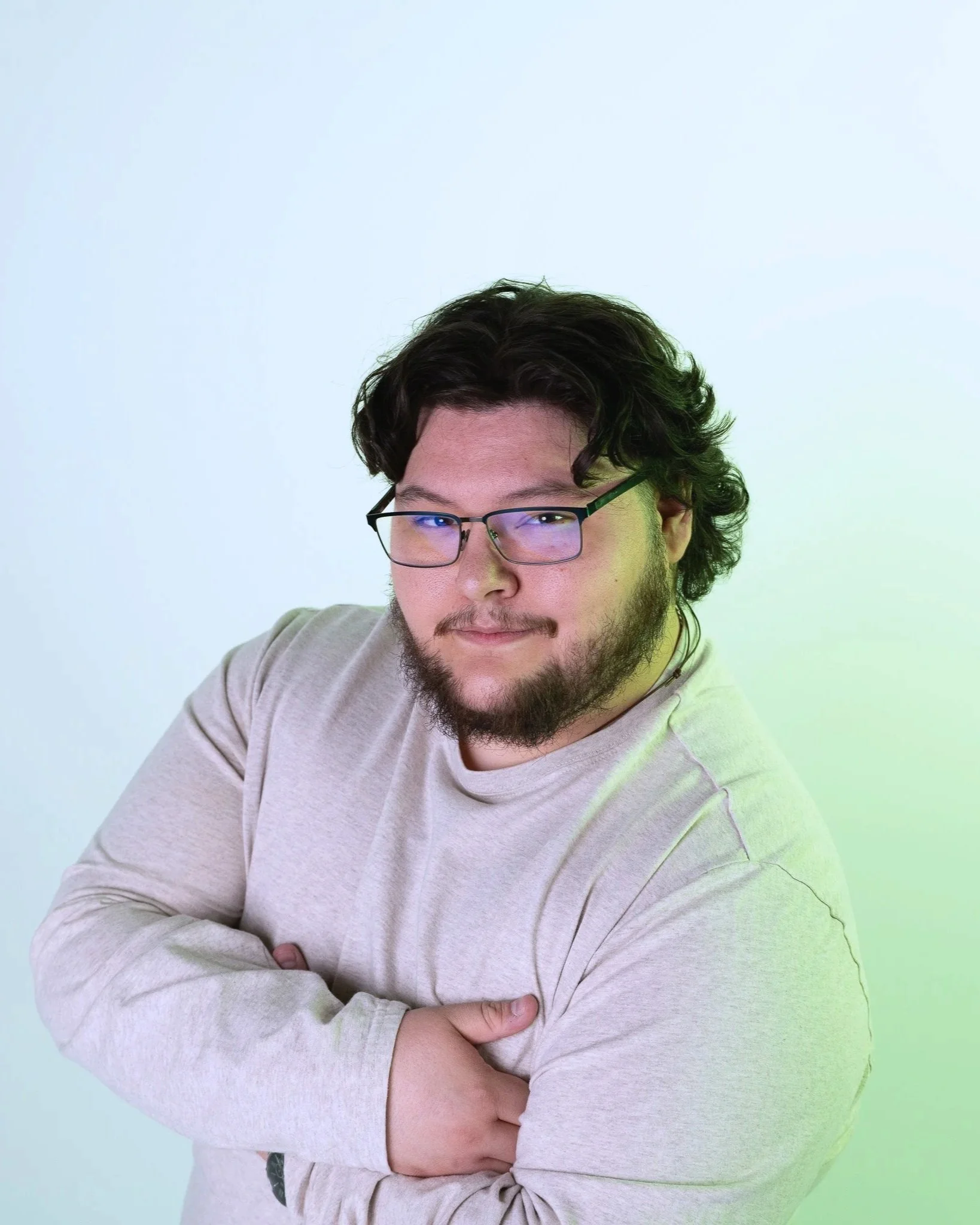 A young man with dark, curly hair and glasses, wearing a beige long-sleeve shirt, standing against a light green and blue background with crossed arms.