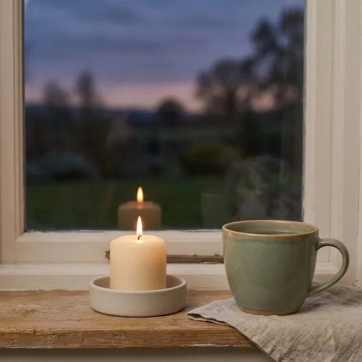 Lit candle on a windowsill with a coffee mug and cloth, overlooking an outdoor scene during dusk.