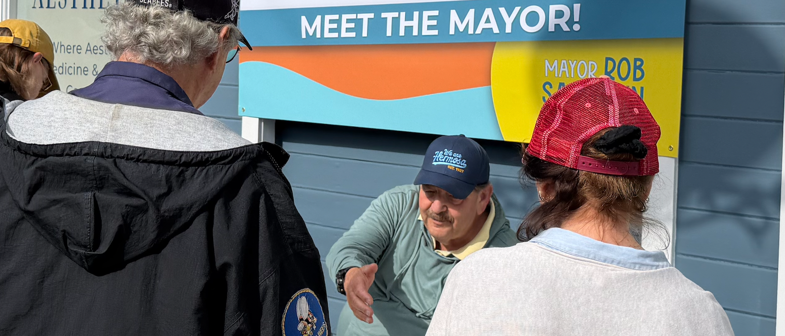 A man wearing a blue cap and light green jacket is sitting at a table, speaking to a small group of people. The group includes a woman with a red cap, a man with curly gray hair and a black jacket, and a woman with brown hair and a yellow cap. They are outdoors, with a large sign behind them that says 'Meet the Mayor!'