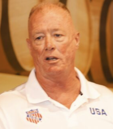 A middle-aged man with red hair and freckles wearing a white polo shirt with USA and a logo on it, standing in front of barrels.