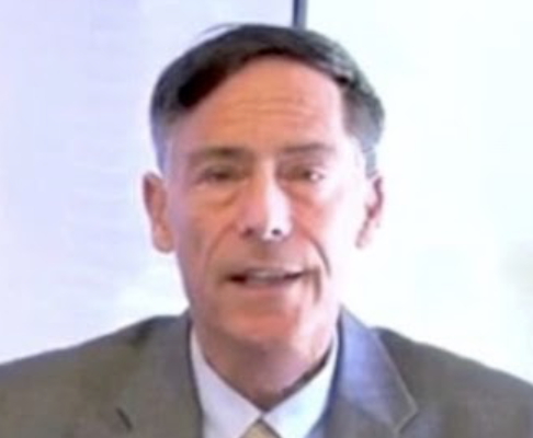 A middle-aged man with dark hair, wearing a gray suit and white shirt, speaking or presenting in front of a light background.