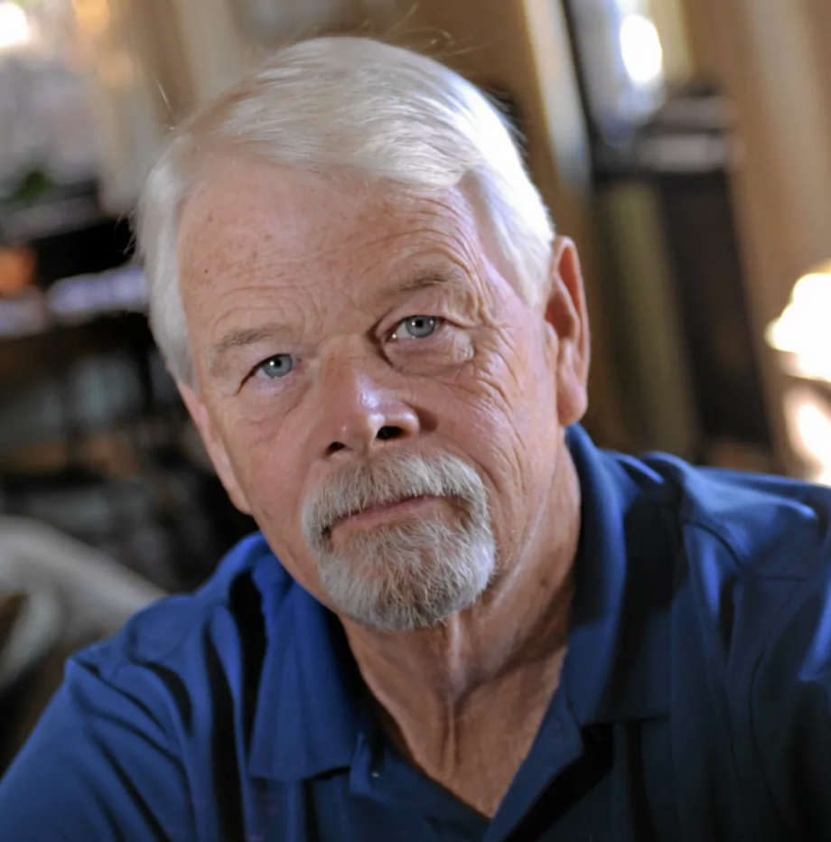 Close-up of an elderly man with white hair and a beard, wearing a blue shirt, in a warmly lit room.