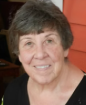 A smiling older woman with short gray hair, wearing a black top, standing in front of an orange background.