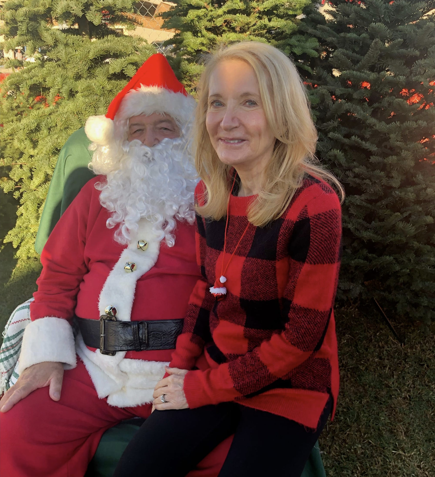 A woman with blonde hair in a red and black plaid sweater sitting next to a man dressed as Santa Claus with a red suit and hat, white beard, and a green cape, outdoors in front of a Christmas tree.
