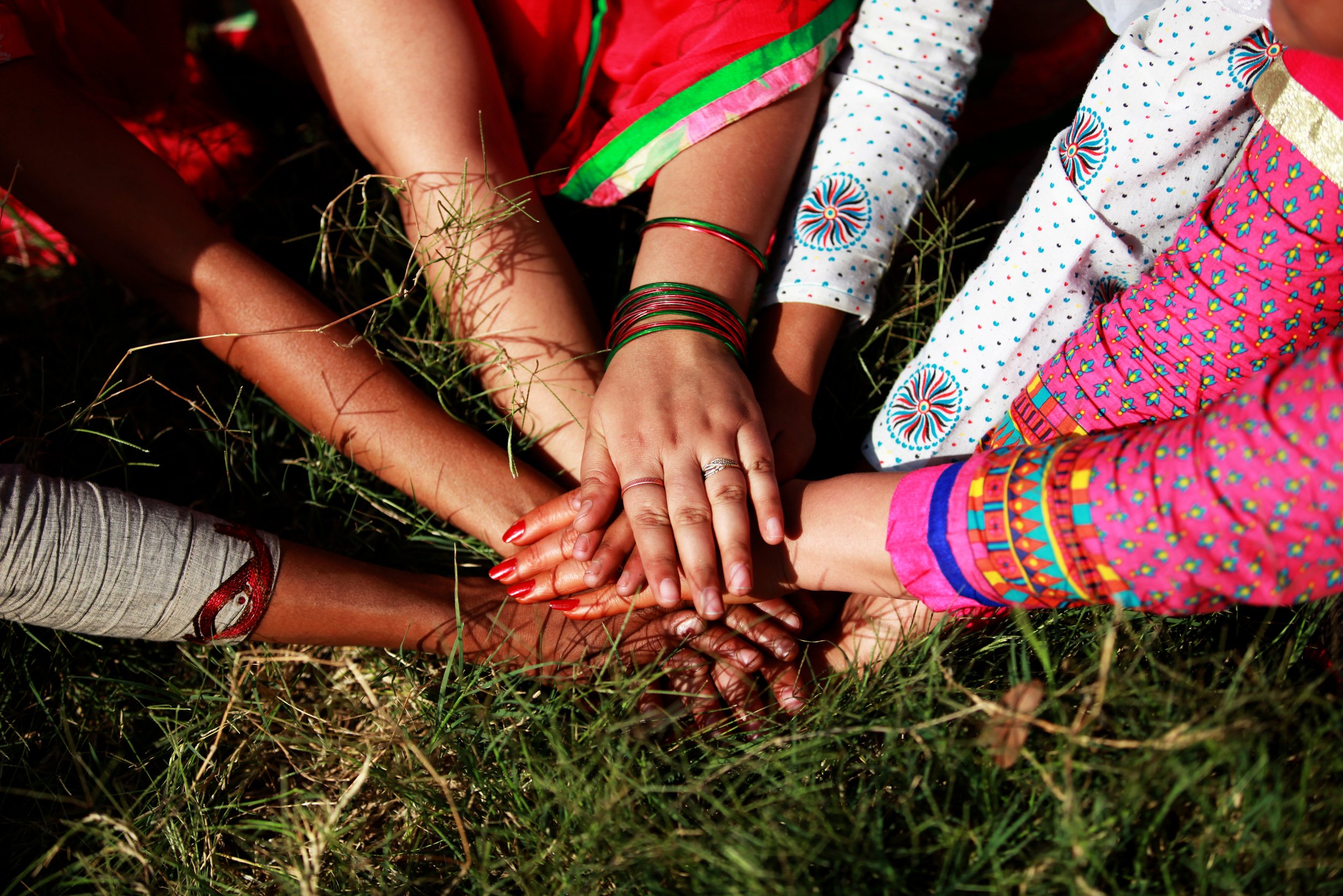 Multiple people's hands stacked together on the grass, some wearing traditional clothing and jewelry, symbolizing unity or teamwork.