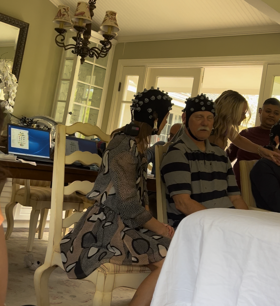 People gathered around a dining table, some wearing black hats with white decorations, in a bright, cozy room with large windows, a chandelier, and laptops on the table.