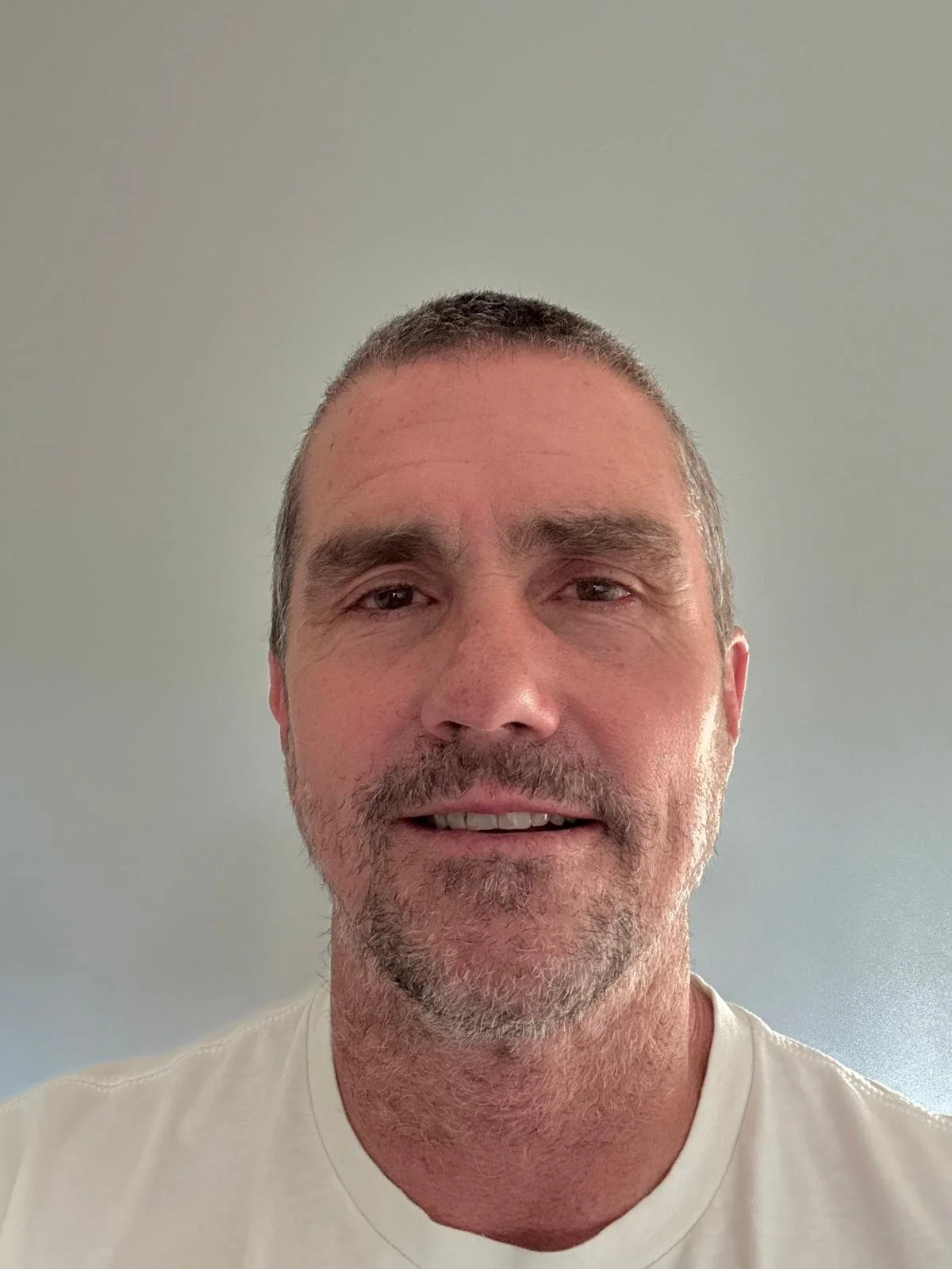 A middle-aged man with short gray hair and facial hair smiling while wearing a white shirt, standing against a plain light green wall.