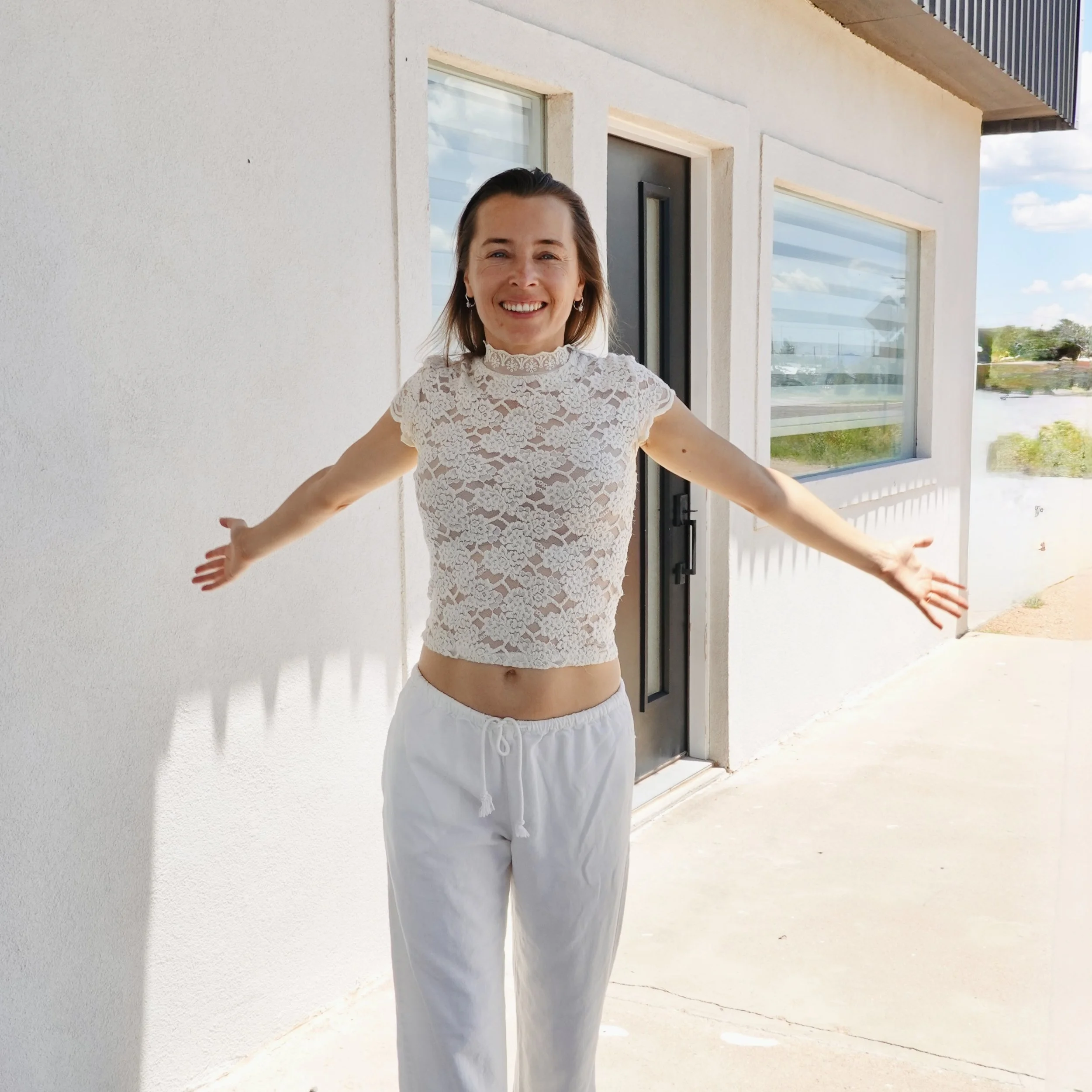 Smiling woman standing outside near a modern house with large windows, wearing a lace top and white pants, with her arms outstretched.