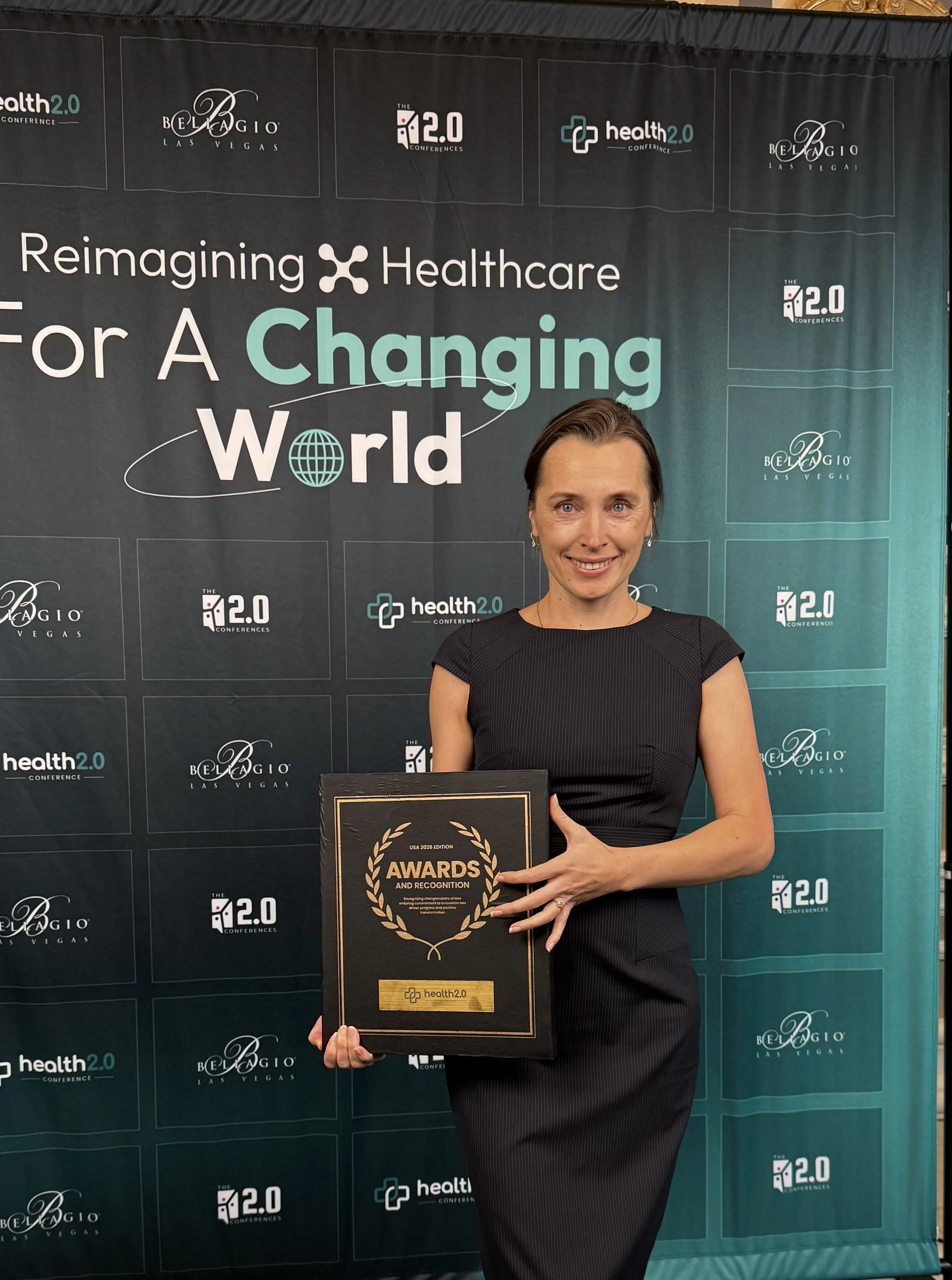 A woman in a black dress holding an award plaque in front of a black backdrop with logos and text related to a healthcare conference. The plaque reads 'AWARDS AND RECOGNITION' and features the 'health2.0' logo.