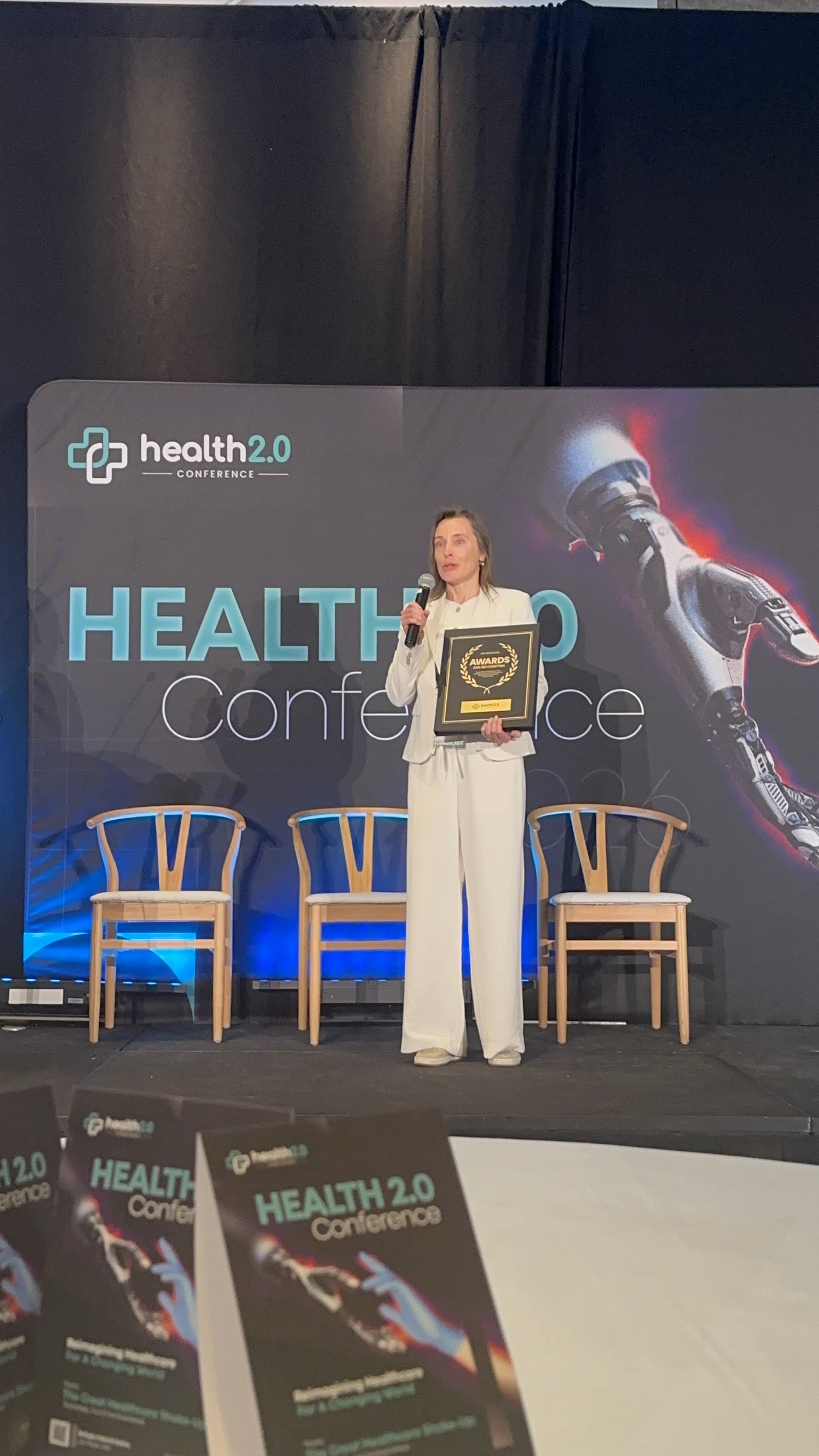 A woman in white suit holding an award and a microphone on stage at a health conference, with chairs in the background and conference branding on the screen and flyers in the foreground.