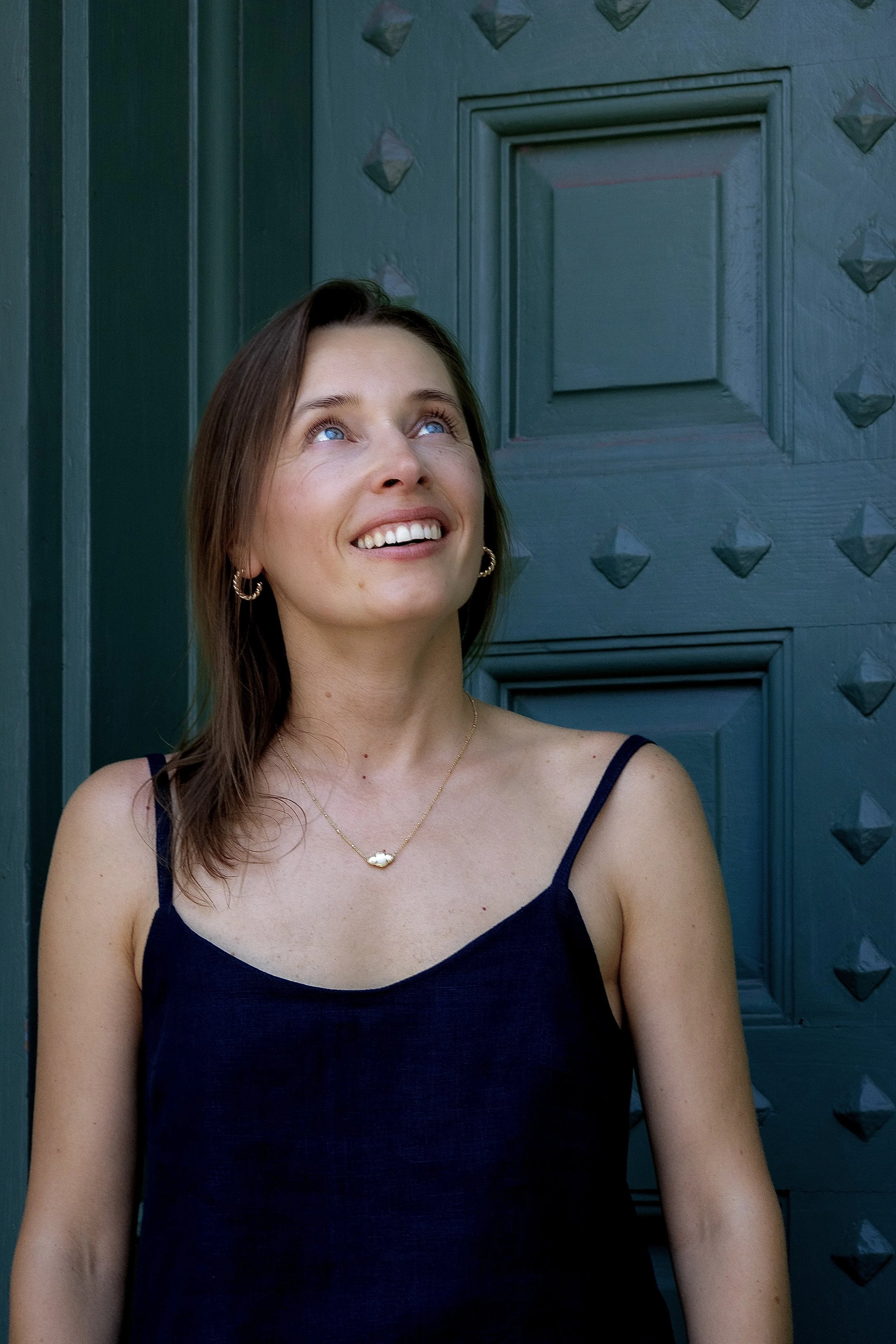 A woman with brown hair and blue eyes smiling, wearing a black sleeveless top, gold hoop earrings, and a gold necklace with a small pendant, standing in front of a blue decorative wooden door with diamond-shaped studs.