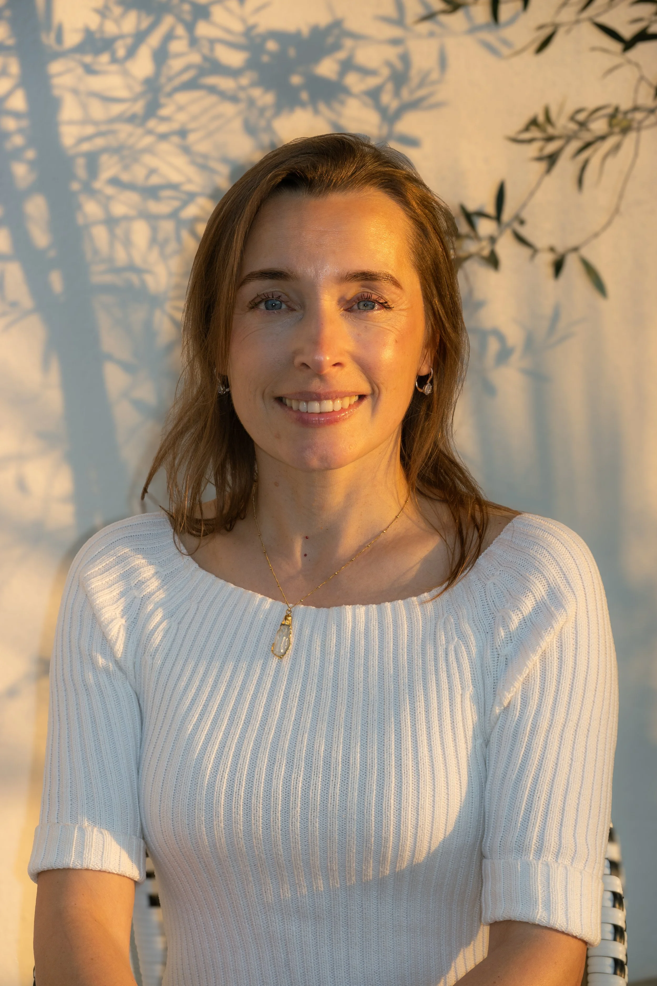 A woman with light brown hair, blue eyes, and fair skin is smiling and wearing a white, ribbed, short-sleeve top with the sleeves rolled up. She has a gold necklace with a pendant and earrings. The background shows a beige wall with shadows of tree branches and leaves.