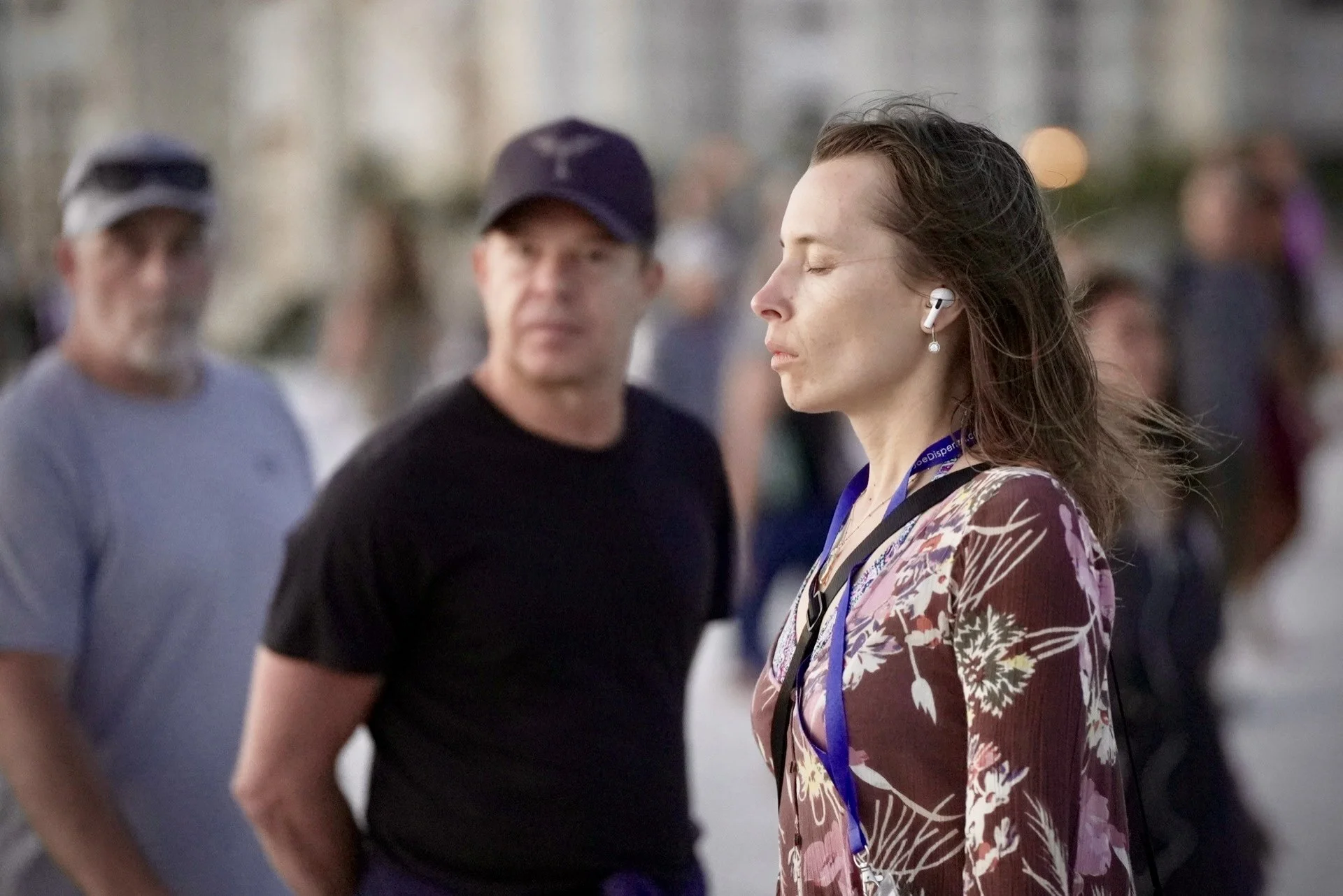 A woman with closed eyes, wearing a floral dress, earbuds, and a lanyard, standing outdoors among blurred people.
