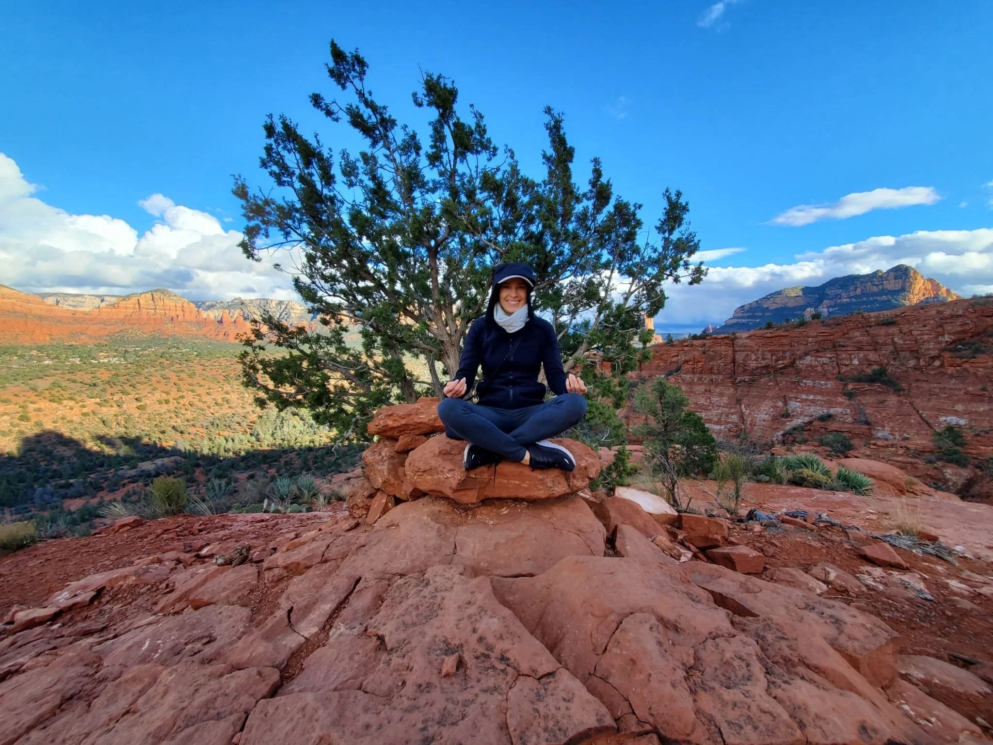 Person meditating on red rocks in a desert landscape with a large tree, mountains, and a blue sky in the background.