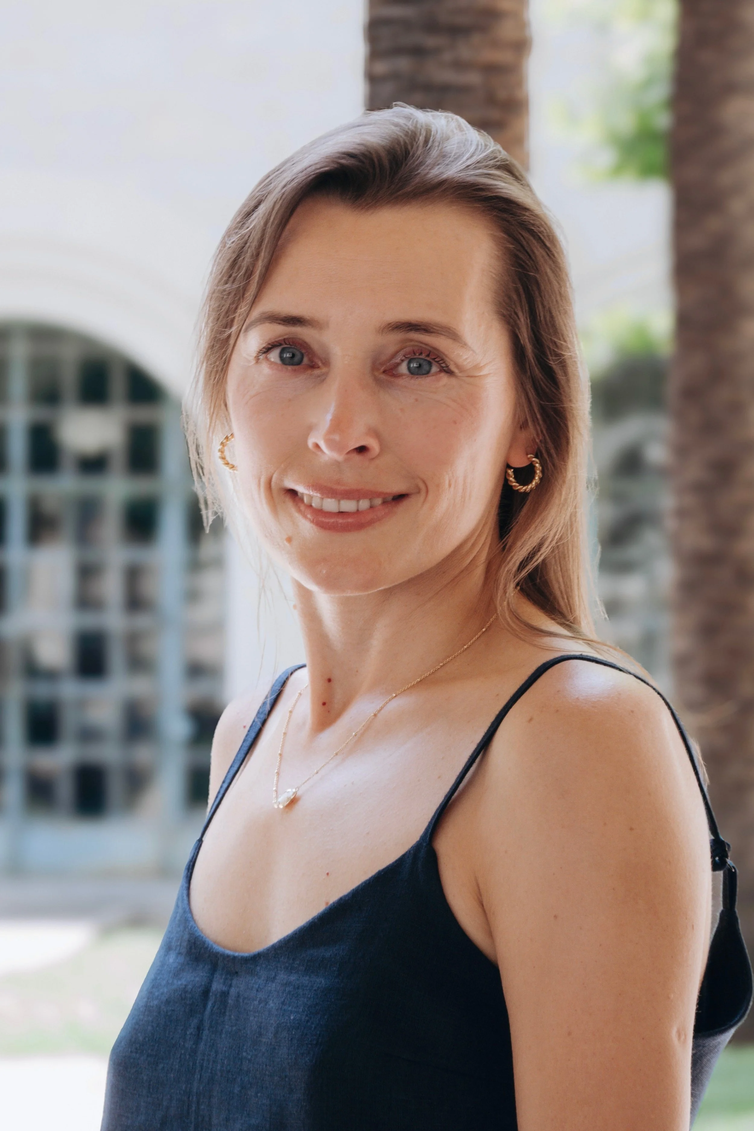 Close-up of a woman with blonde hair, blue eyes, and fair skin, smiling slightly, wearing a black spaghetti strap dress, gold hoop earrings, and layered necklaces, with a blurred background of a building and greenery.