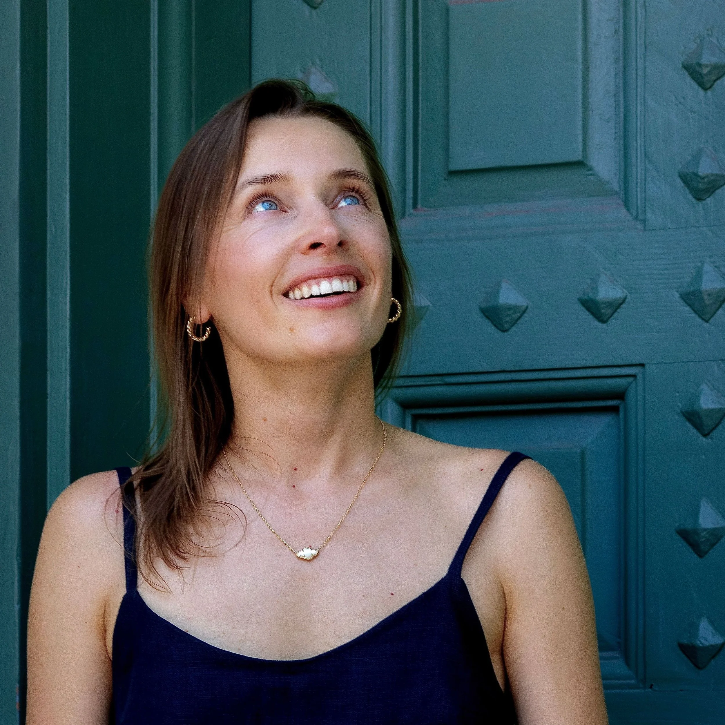 Close-up of a woman smiling and looking up, wearing a navy blue tank top, gold jewelry, and standing in front of a blue-green wooden door.